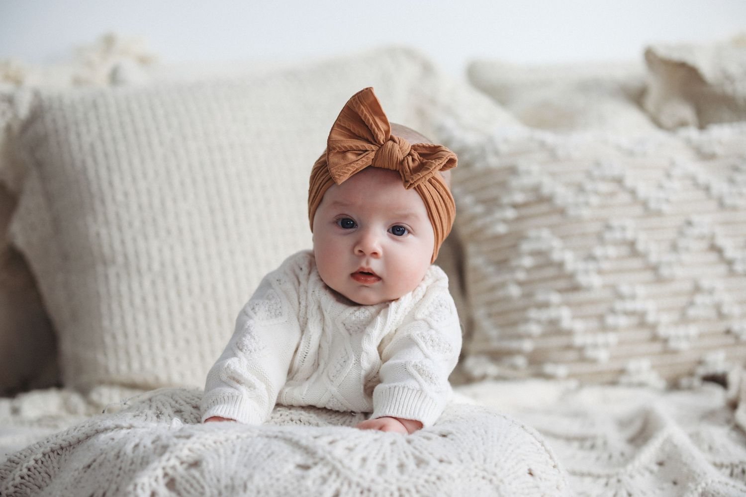 Baby wearing a brown bow headband lies on white textured bedding with decorative pillows in background.