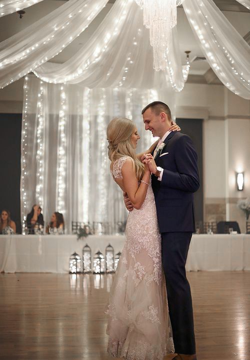 A couple shares an emotional dance in an elegant ballroom decorated with twinkling lights and white fabric draping.