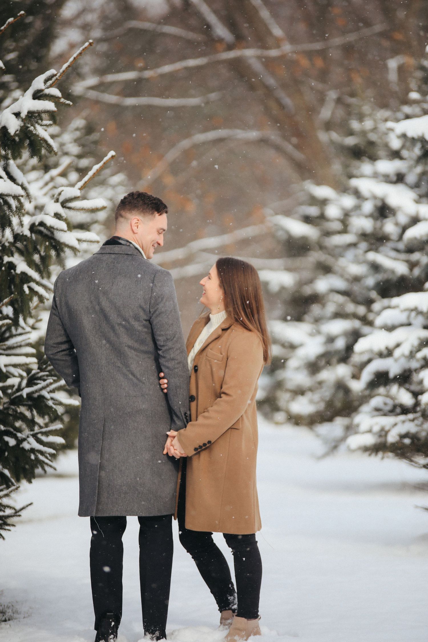 A couple shares a romantic moment in a snowy winter forest setting wearing coats.