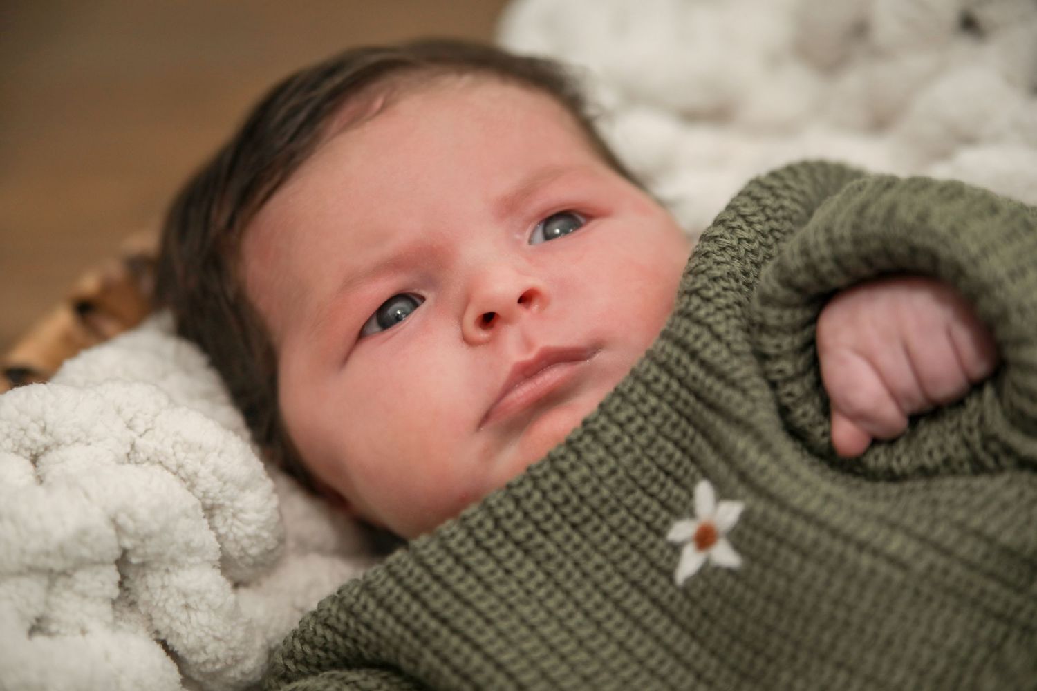 Newborn baby wearing a green knit sweater lying on soft white bedding in a cozy setting.