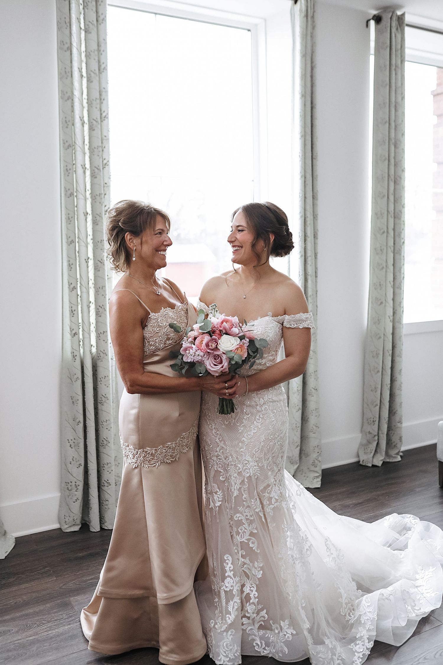 Two women share a joyful moment in formal attire, with one wearing a white wedding gown and the other in champagne-colored dress.