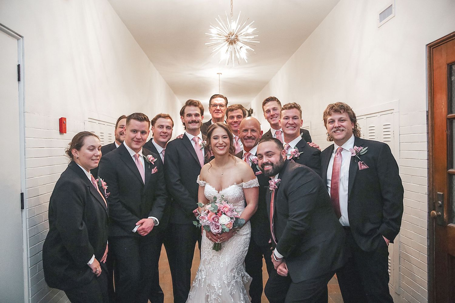Wedding party lines up along a white hallway wall with lockers for a casual group photo.