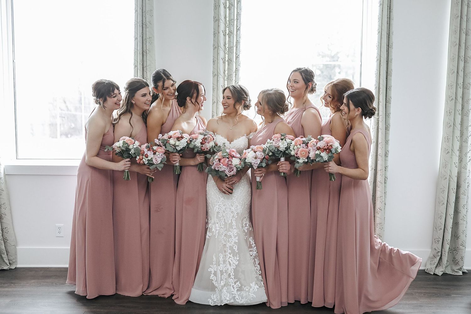 Bridesmaids in dusty rose dresses stand together with bouquets in an elegant white room during a wedding celebration.