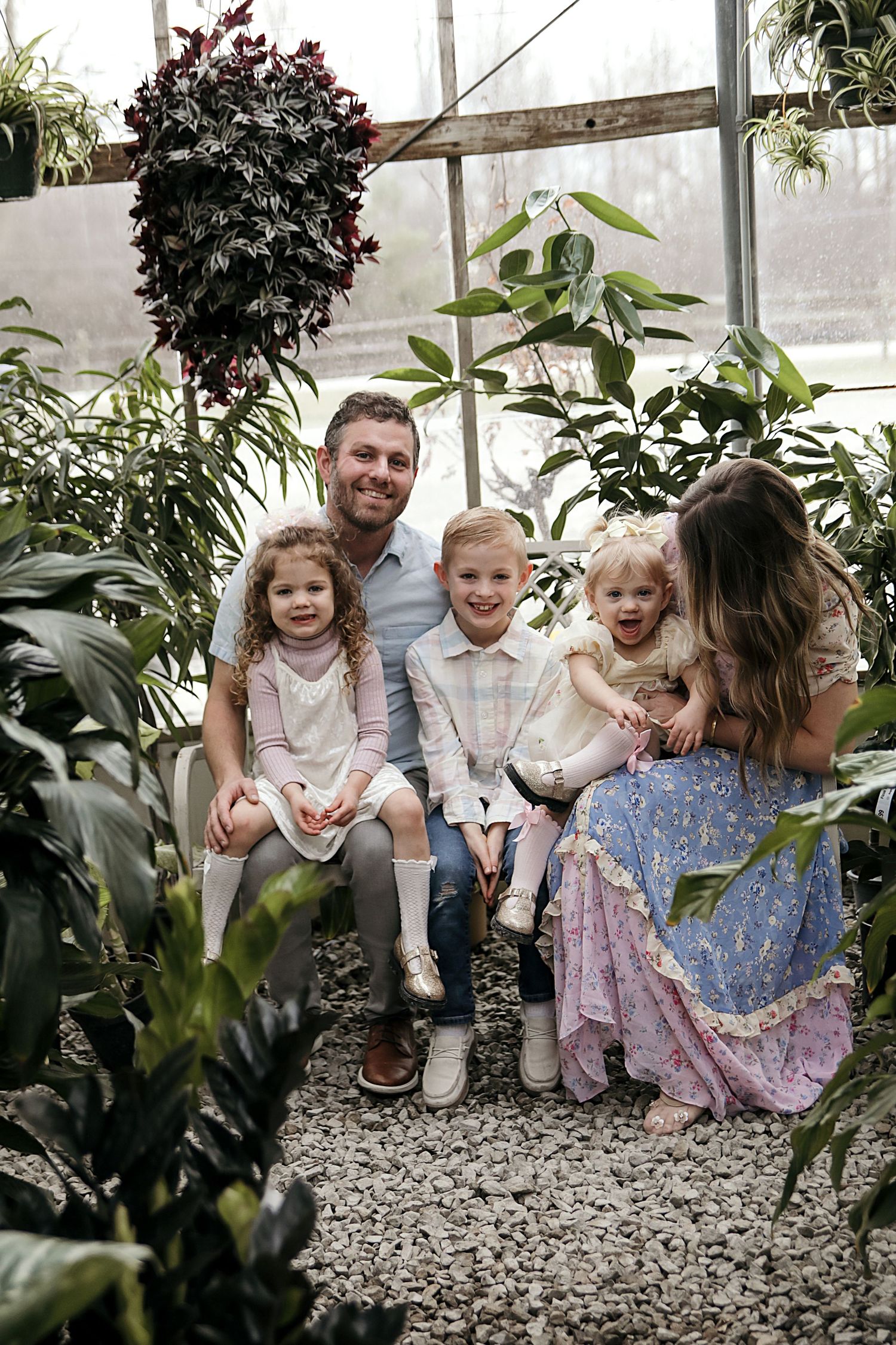 A family sits together surrounded by lush plants and greenery in a glassed-in greenhouse space.