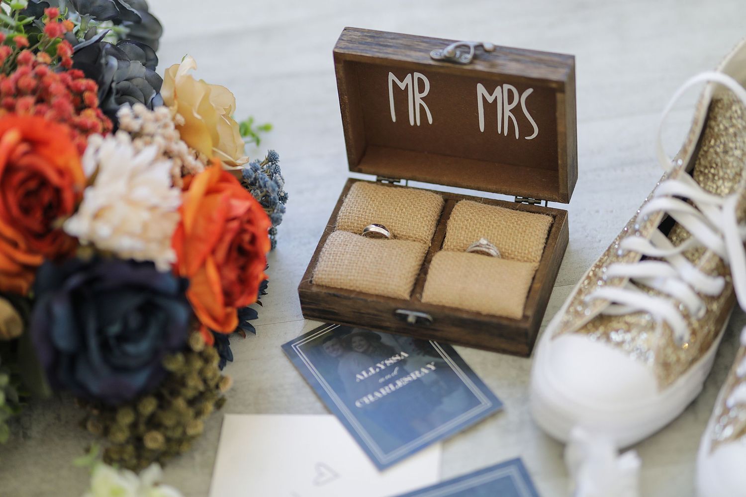 Wedding details with mr mrs wooden ring box, bouquet of orange and blue flowers, and white lace shoes on display.