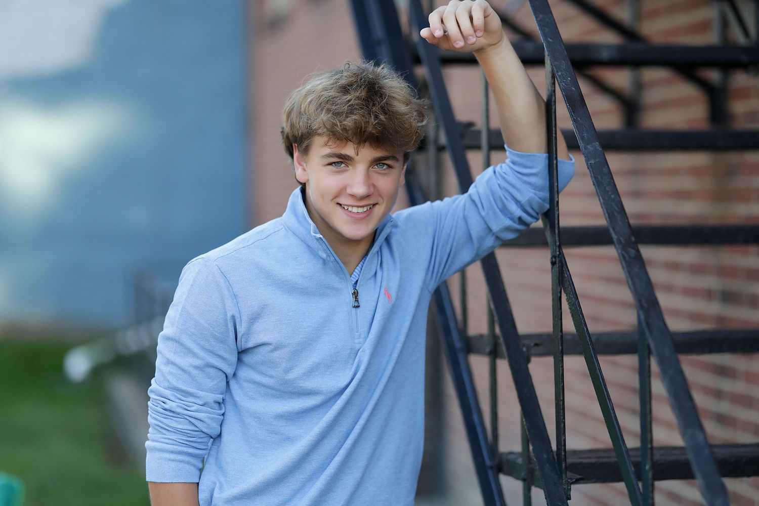Young person in a light blue sweater gesturing and posing against a brick wall background.