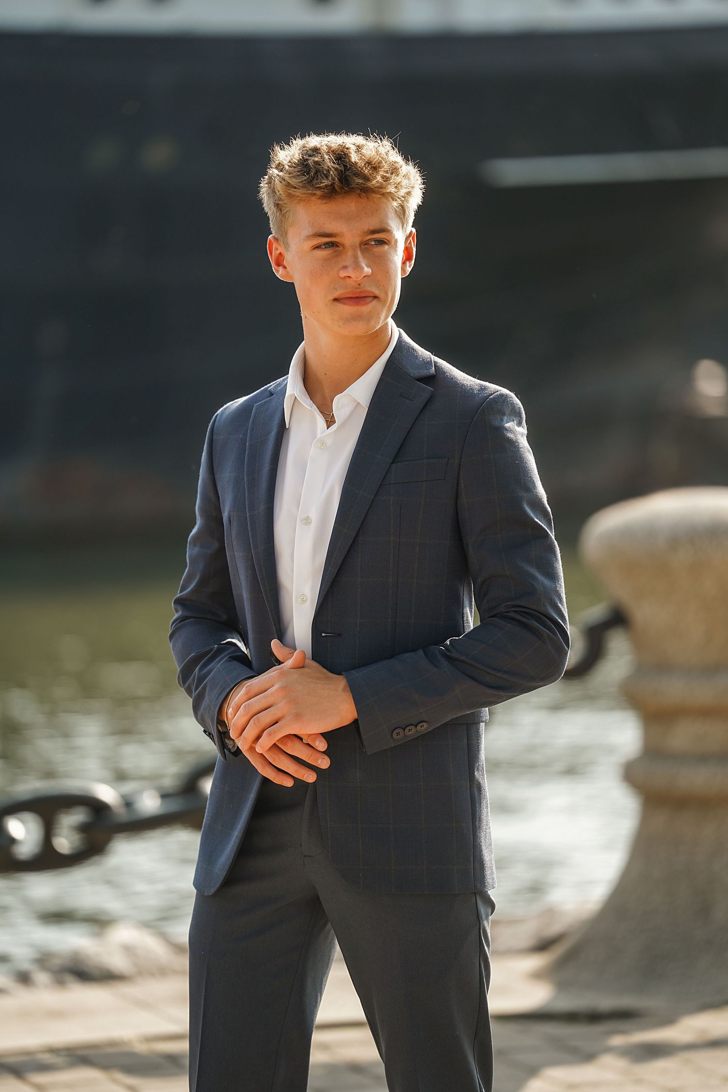 Professional portrait of a person wearing a navy suit jacket and white shirt standing by a waterfront in sunlight.