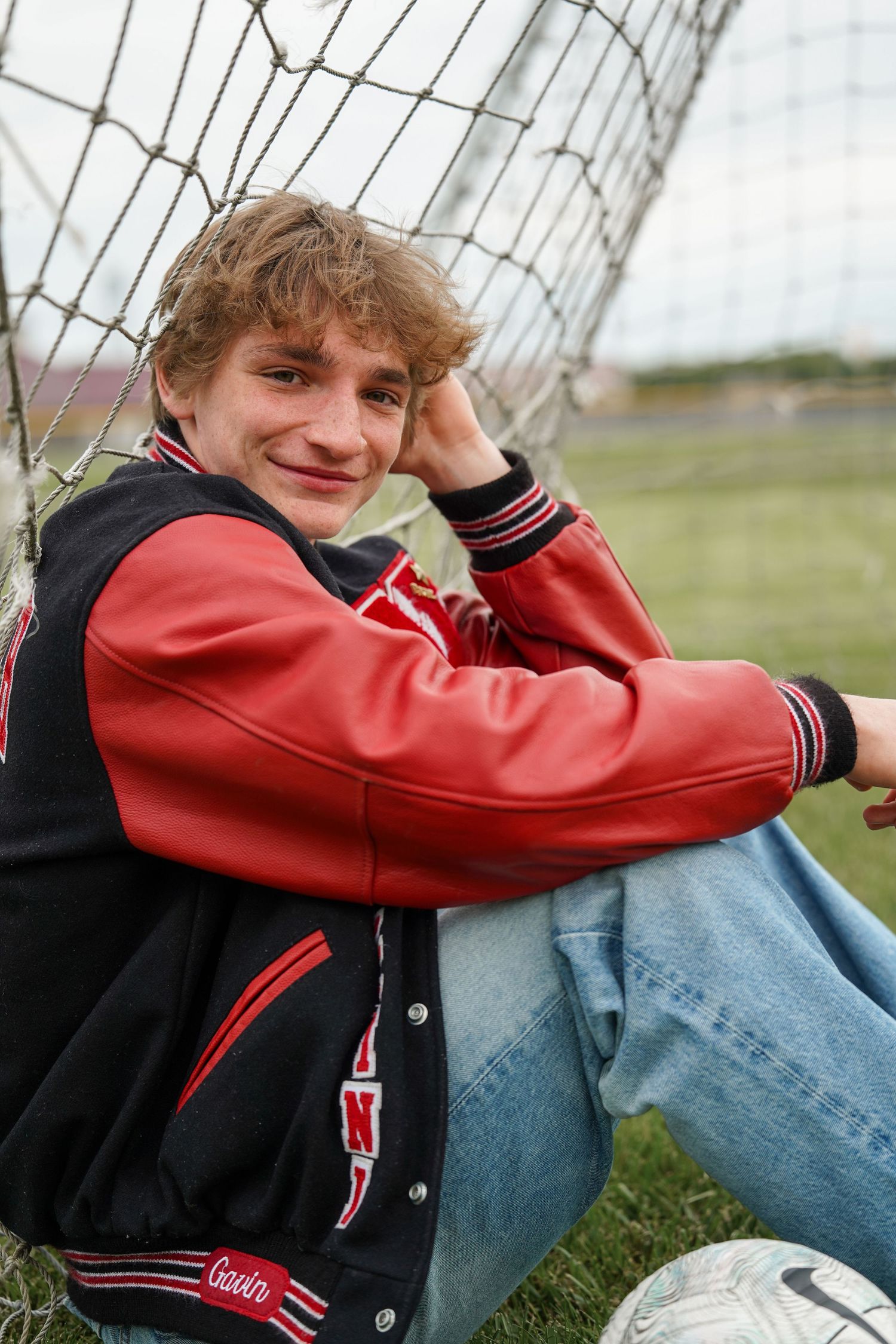 Student athlete in red and black varsity jacket sits casually against soccer goal net with ball nearby on grass field.