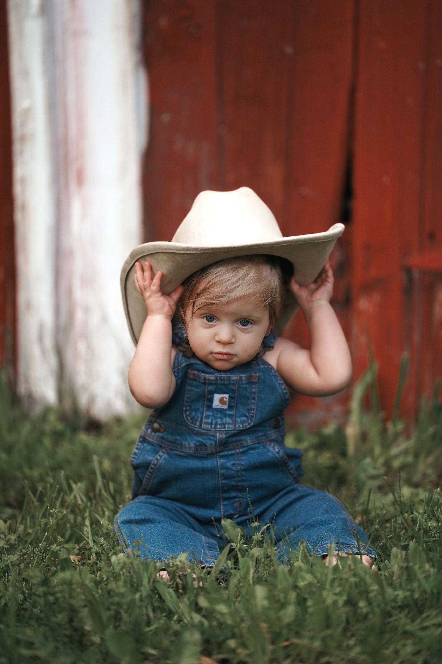 Small child in denim overalls sits in grass wearing an oversized cowboy hat against red barn backdrop.