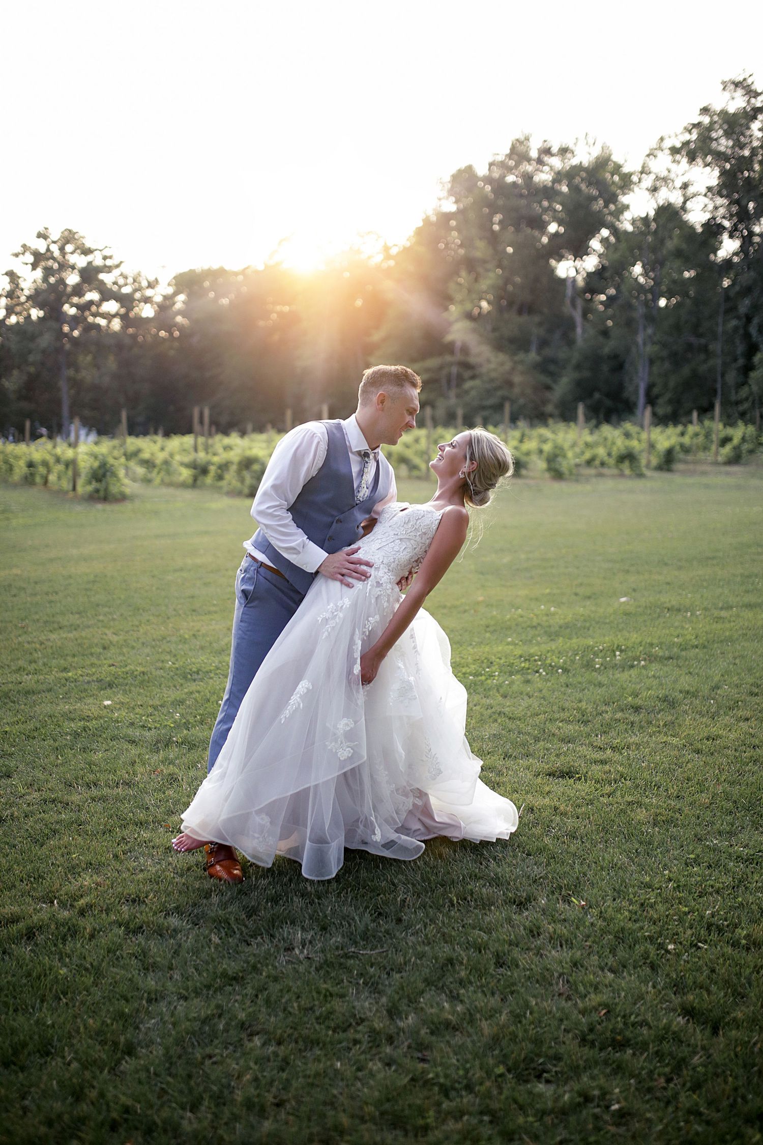 A wedding couple shares a romantic sunset dance in a vineyard field with golden sunlight glowing through trees.