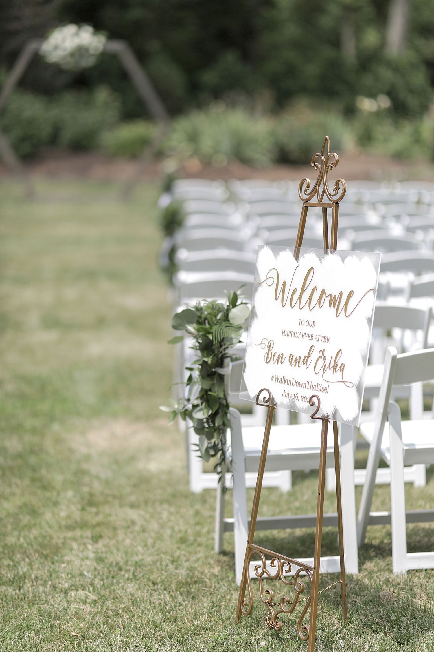 Elegant welcome sign with greenery displayed on a gold easel at an outdoor wedding ceremony setup with white chairs.
