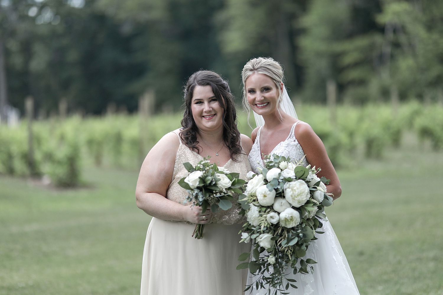 Two bridesmaids holding white floral bouquets pose together in outdoor garden setting.