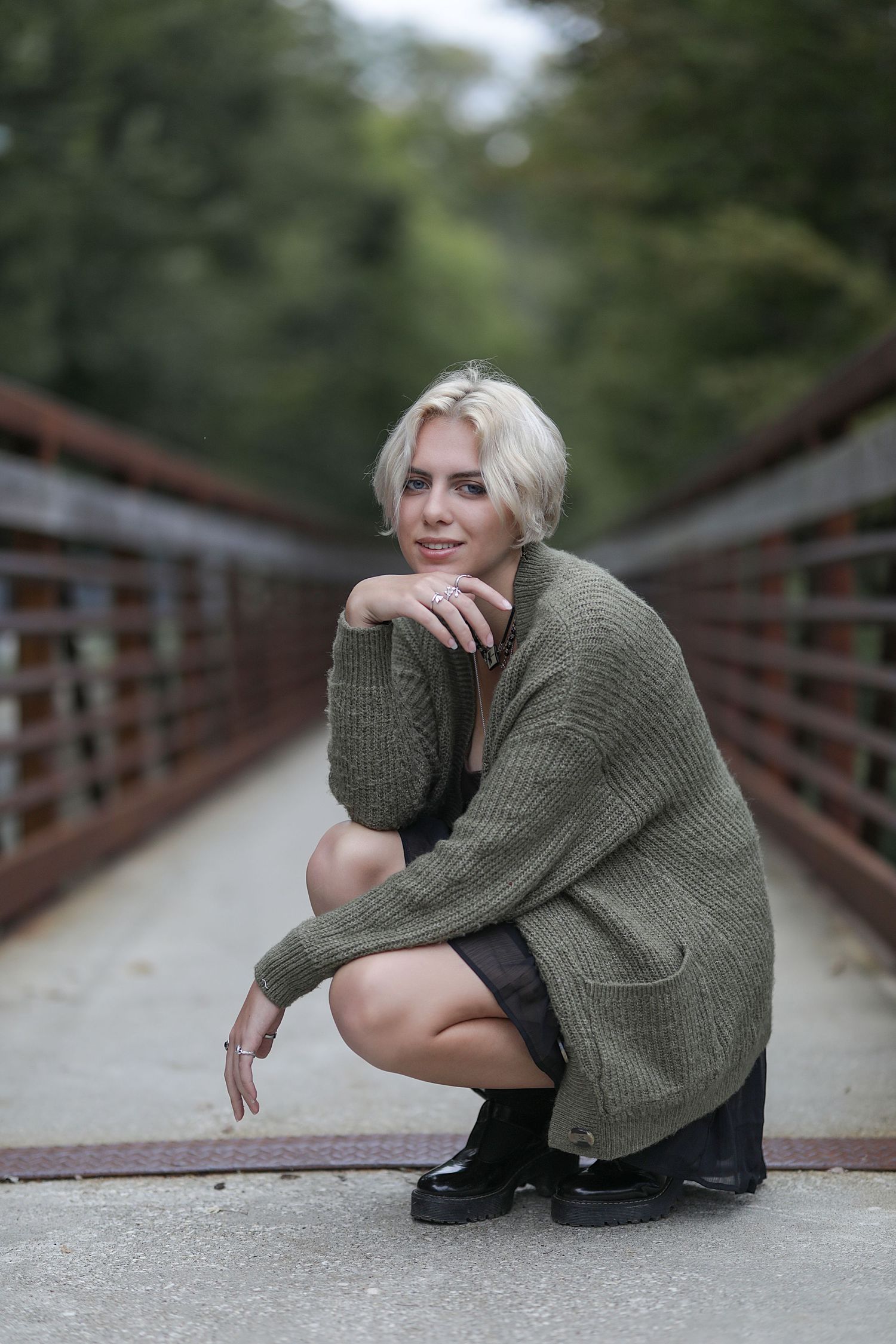Person wearing an oversized olive green sweater crouches on a wooden bridge in a casual outdoor portrait.