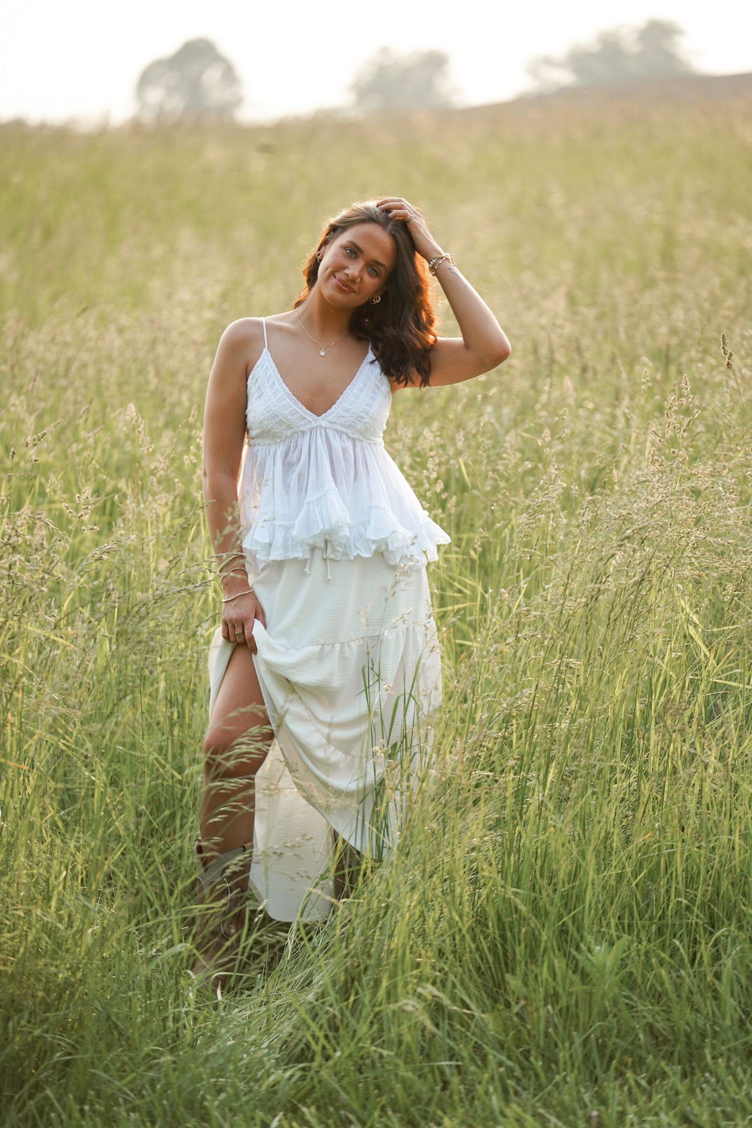 A person in a flowy white dress walks through tall green grass in a field during golden hour sunlight.