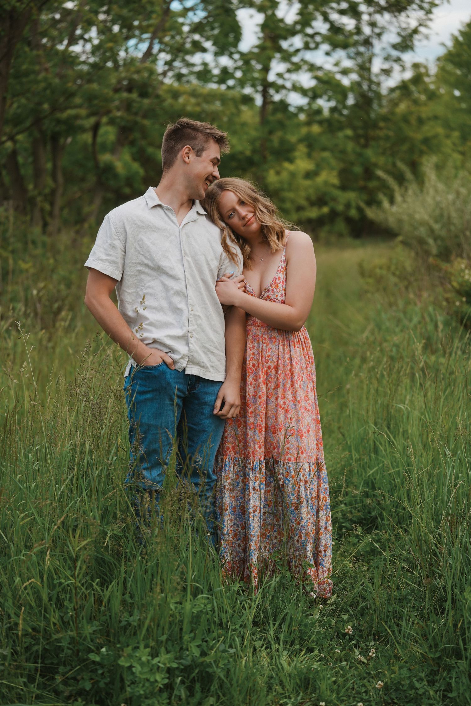A couple stands close together in a green summer meadow filled with tall grass and wild vegetation.