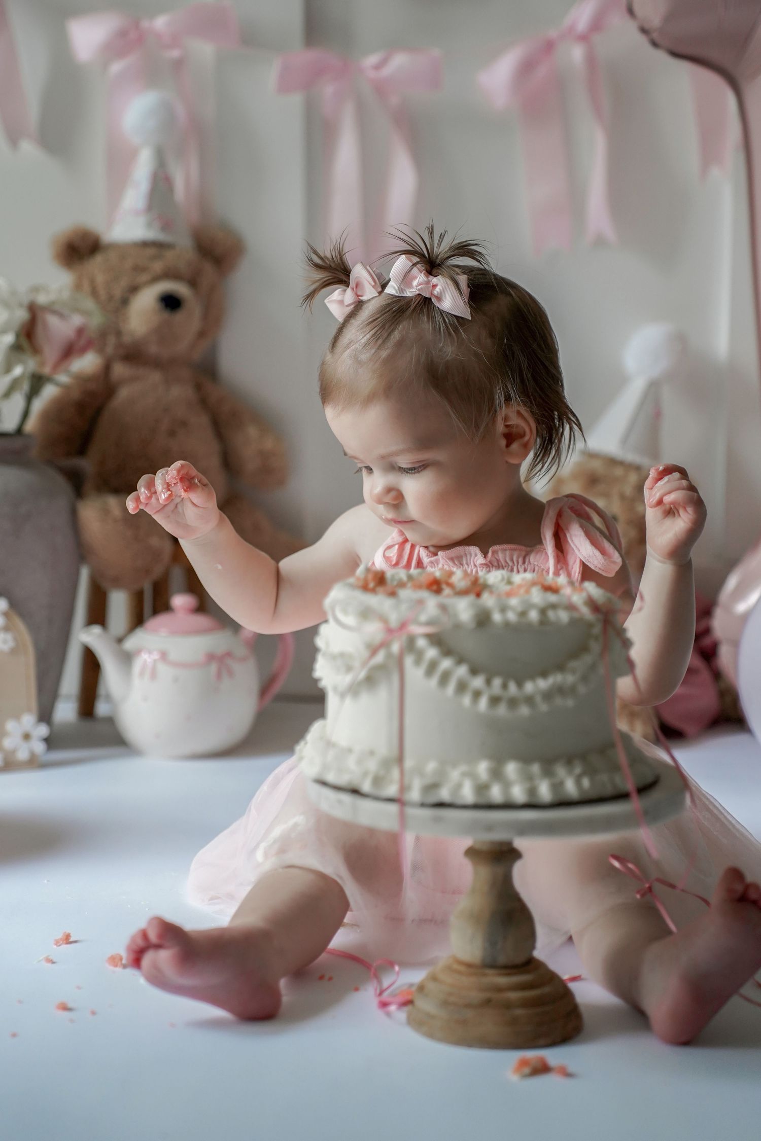 Sweet pink and white themed first birthday cake smash photo with stuffed bear and delicate ribbons in background.