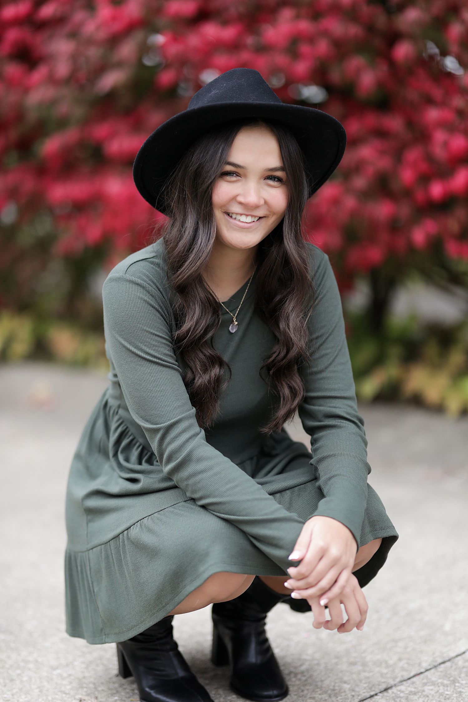 A person in a green sweater dress and black hat crouches down while smiling against a backdrop of pink autumn foliage.