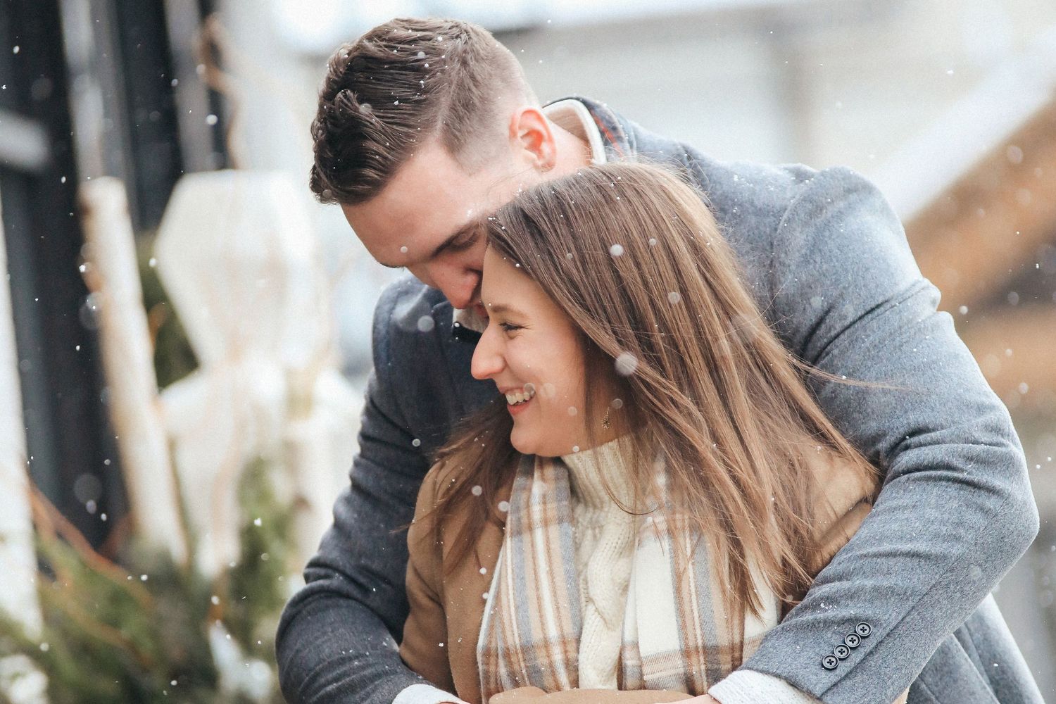 A couple in gray sweaters shares a romantic moment together in falling snow during winter.