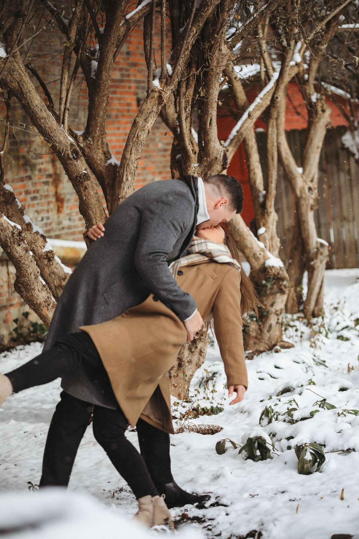 Two people in winter coats dance together playfully in the snow near brick walls and bare trees.