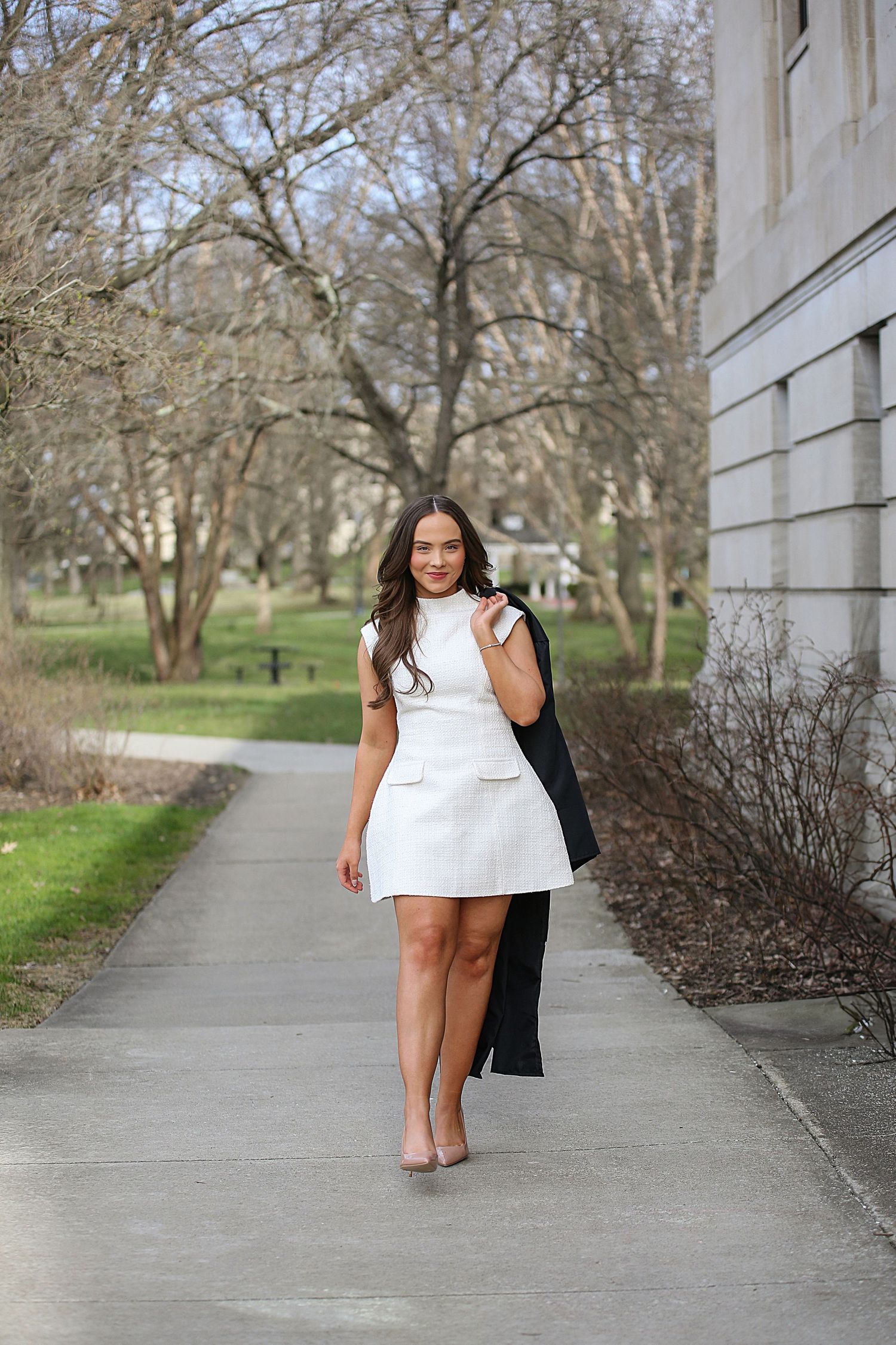 Someone wearing a white sleeveless dress stands on a concrete path surrounded by bare trees and landscaping.