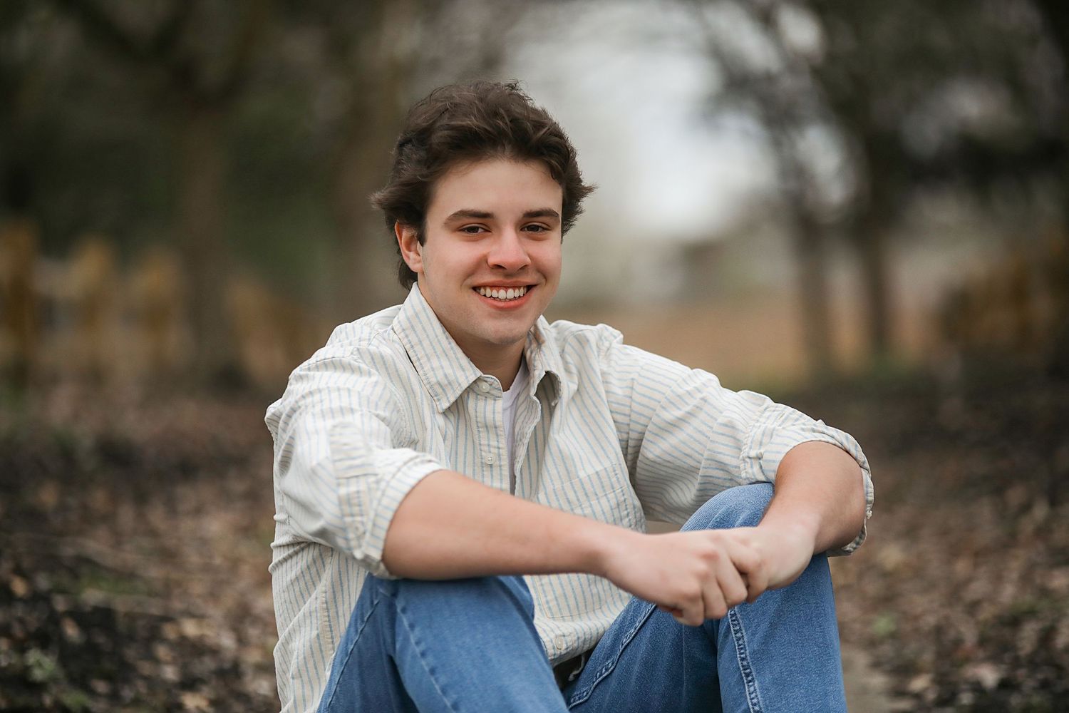 A person in a white button-up shirt and jeans sits casually outdoors during a photo session.