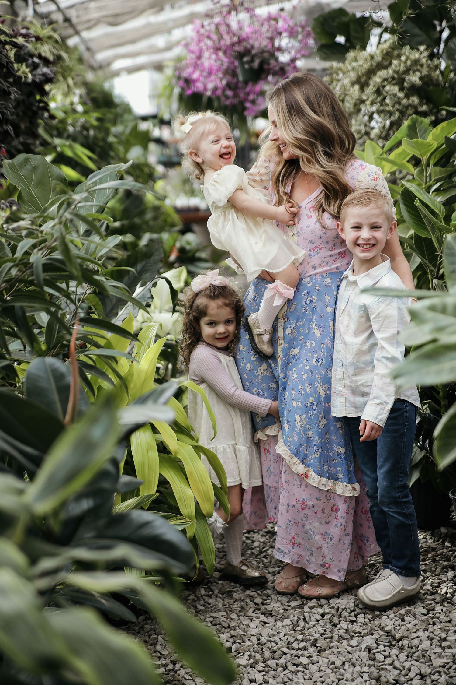 A family shares a joyful moment together inside a lush greenhouse filled with tropical plants and pink flowers.