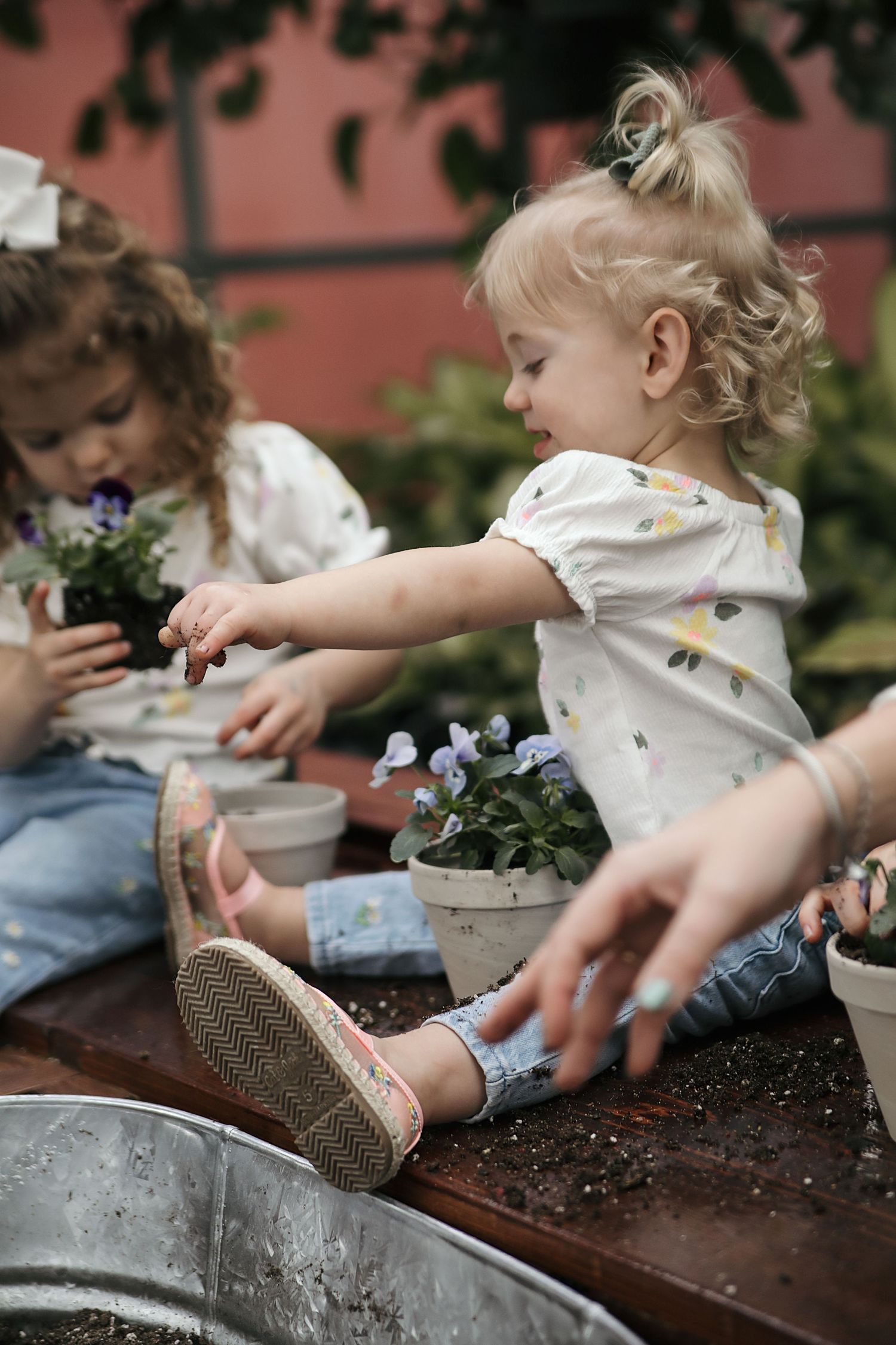 Children planting flowers and playing with soil on a wooden surface near a pink wall, with gardening activities.