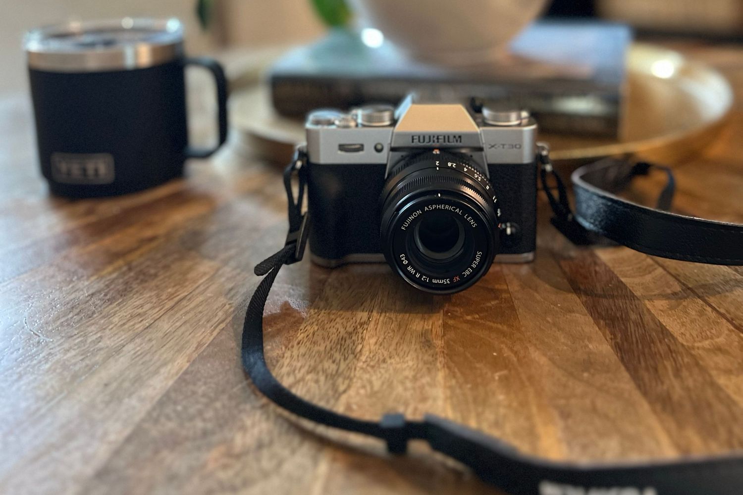 A vintage-style Fujifilm camera with black leather strap sits on a wooden table next to a dark coffee mug.