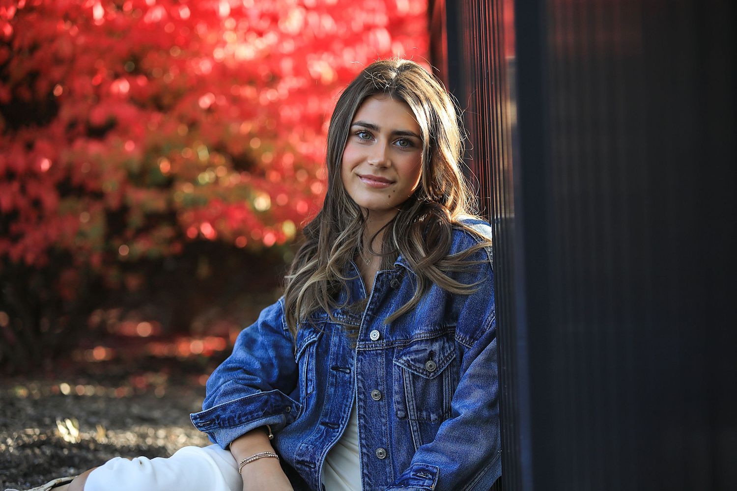 A person in a bright blue jean jacket sits outdoors surrounded by vibrant red autumn foliage.