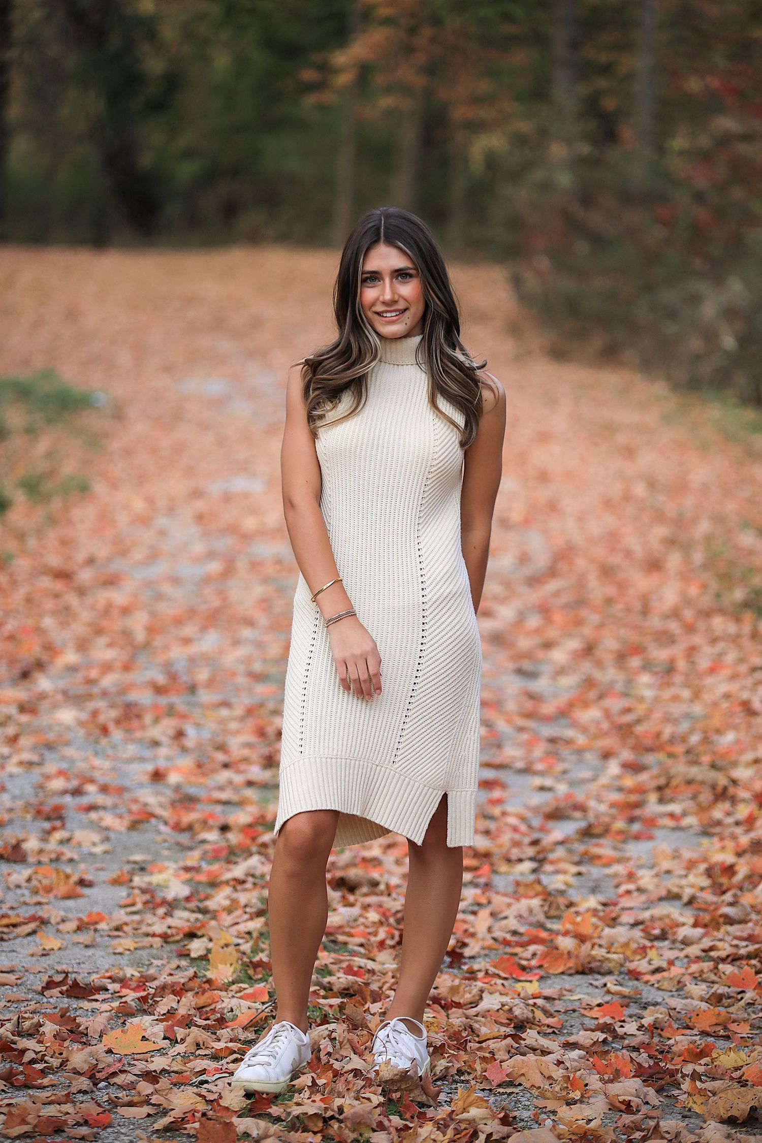 Person in white knit dress poses in autumn leaves wearing white sneakers for a seasonal outdoor photoshoot.