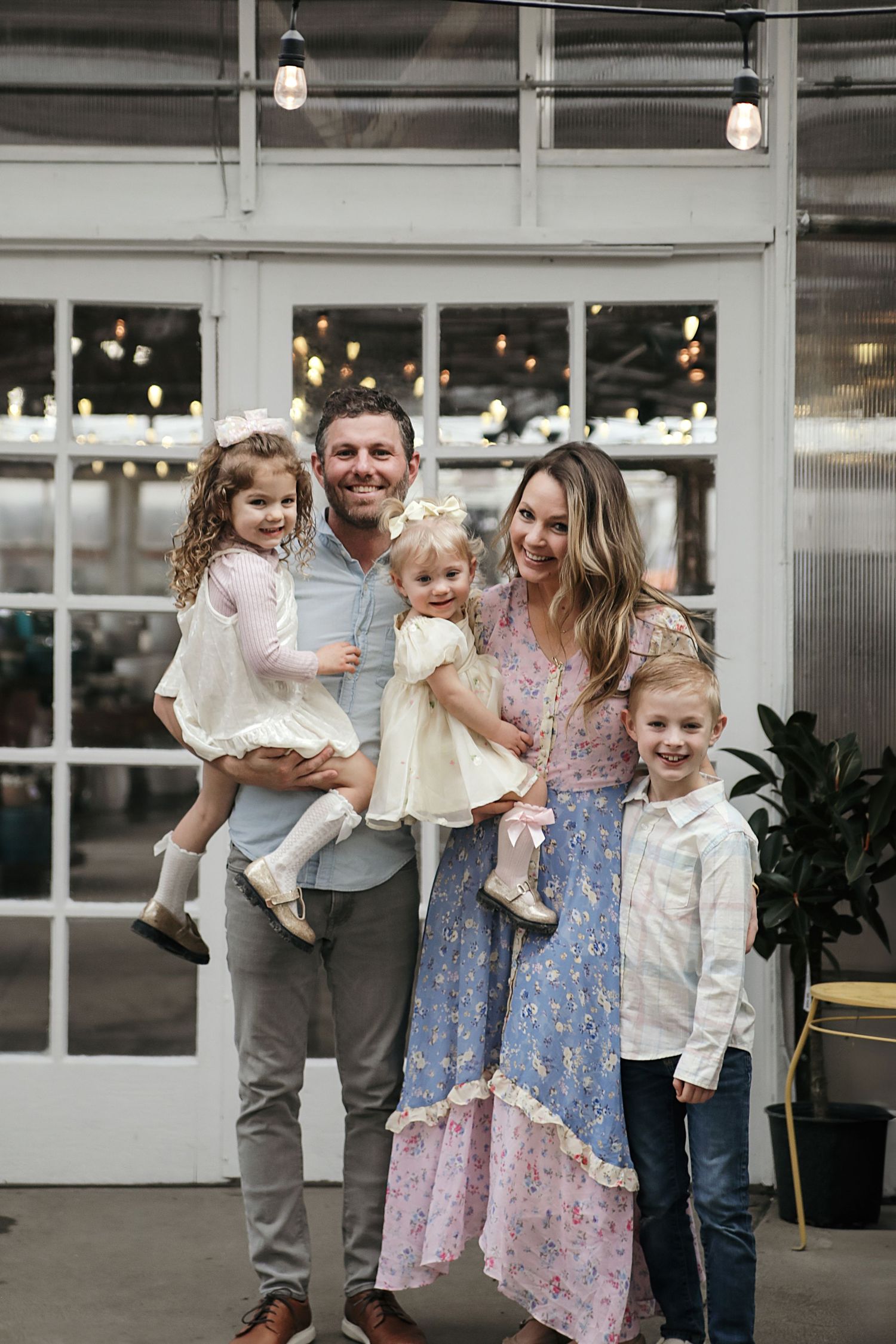 A family poses together inside an industrial-style space with large windows and twinkling lights in the background.