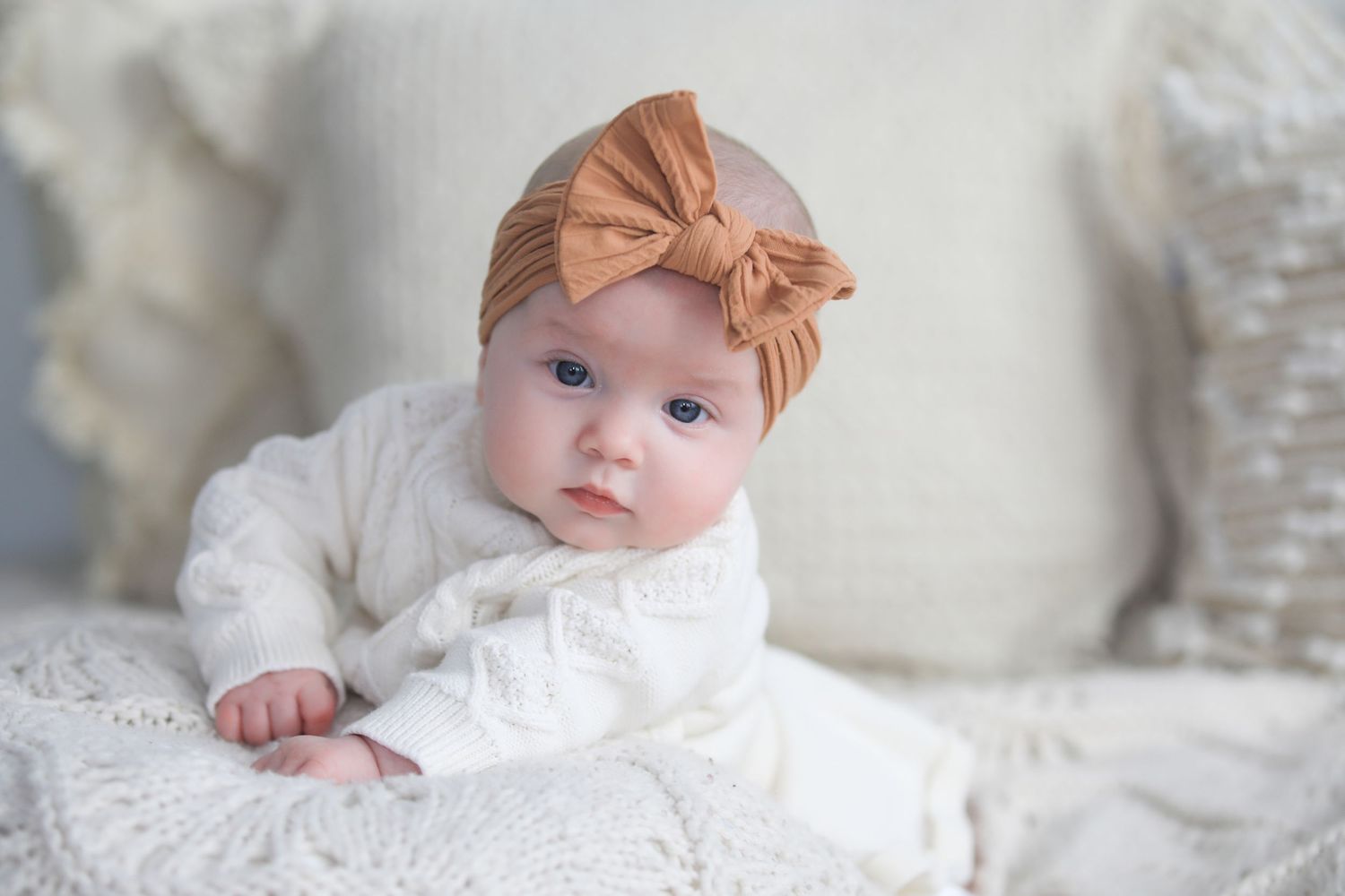 Baby wearing orange bow headband lying on white textured blanket in soft natural lighting.