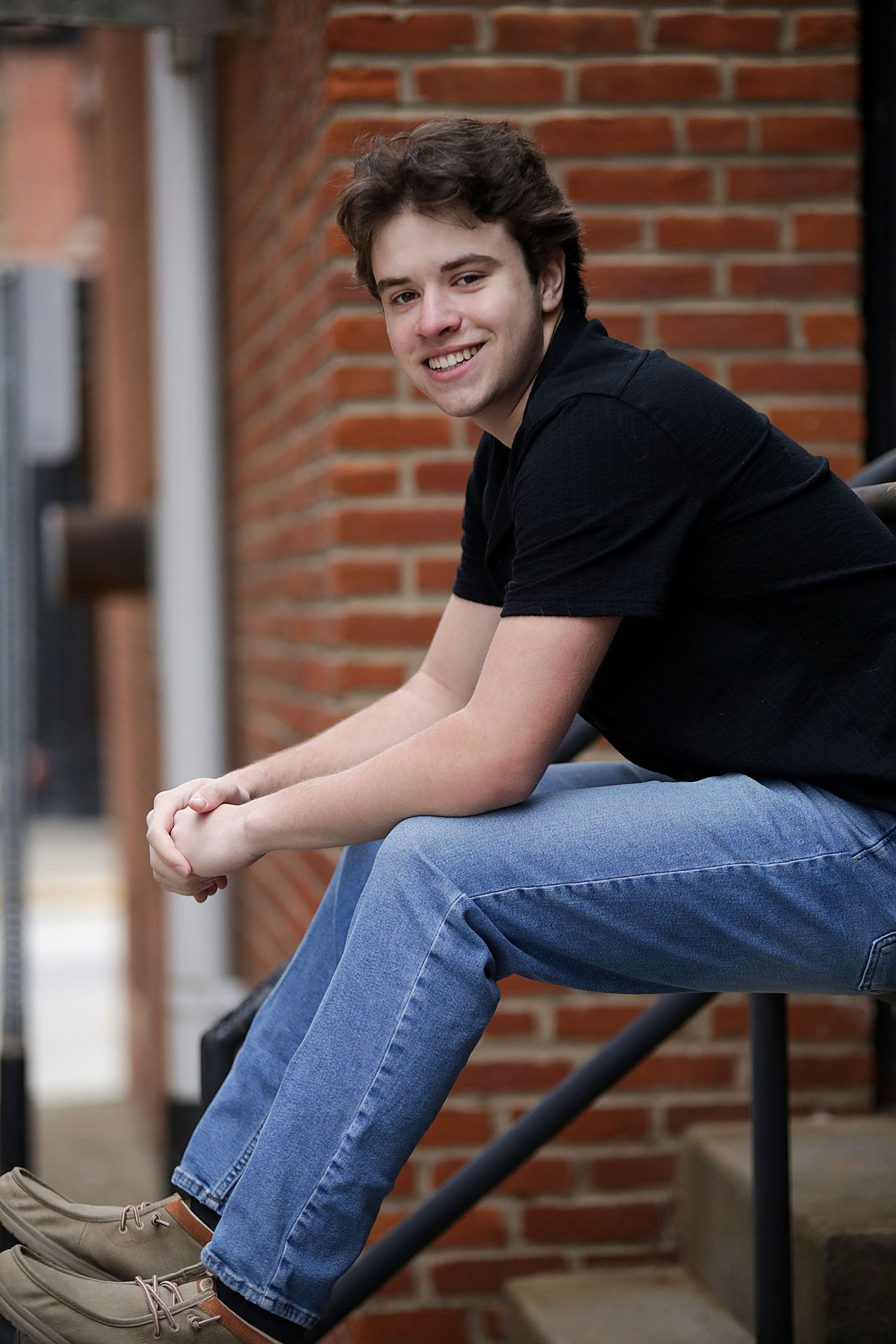 Person in black shirt and jeans sits casually on metal railing against brick wall backdrop.