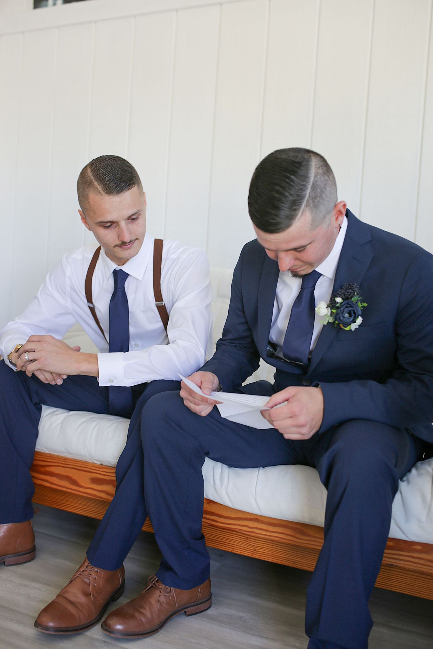 Groomsmen in navy suits and brown shoes sitting on bench looking at something together.