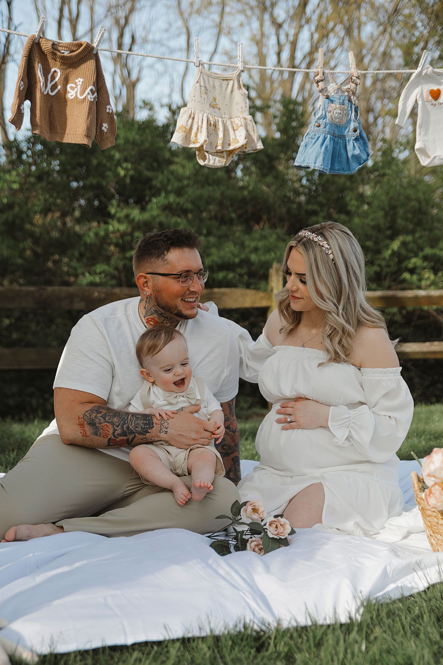 A couple sits on a picnic blanket outdoors with baby clothing hanging on a clothesline behind them in a garden setting.