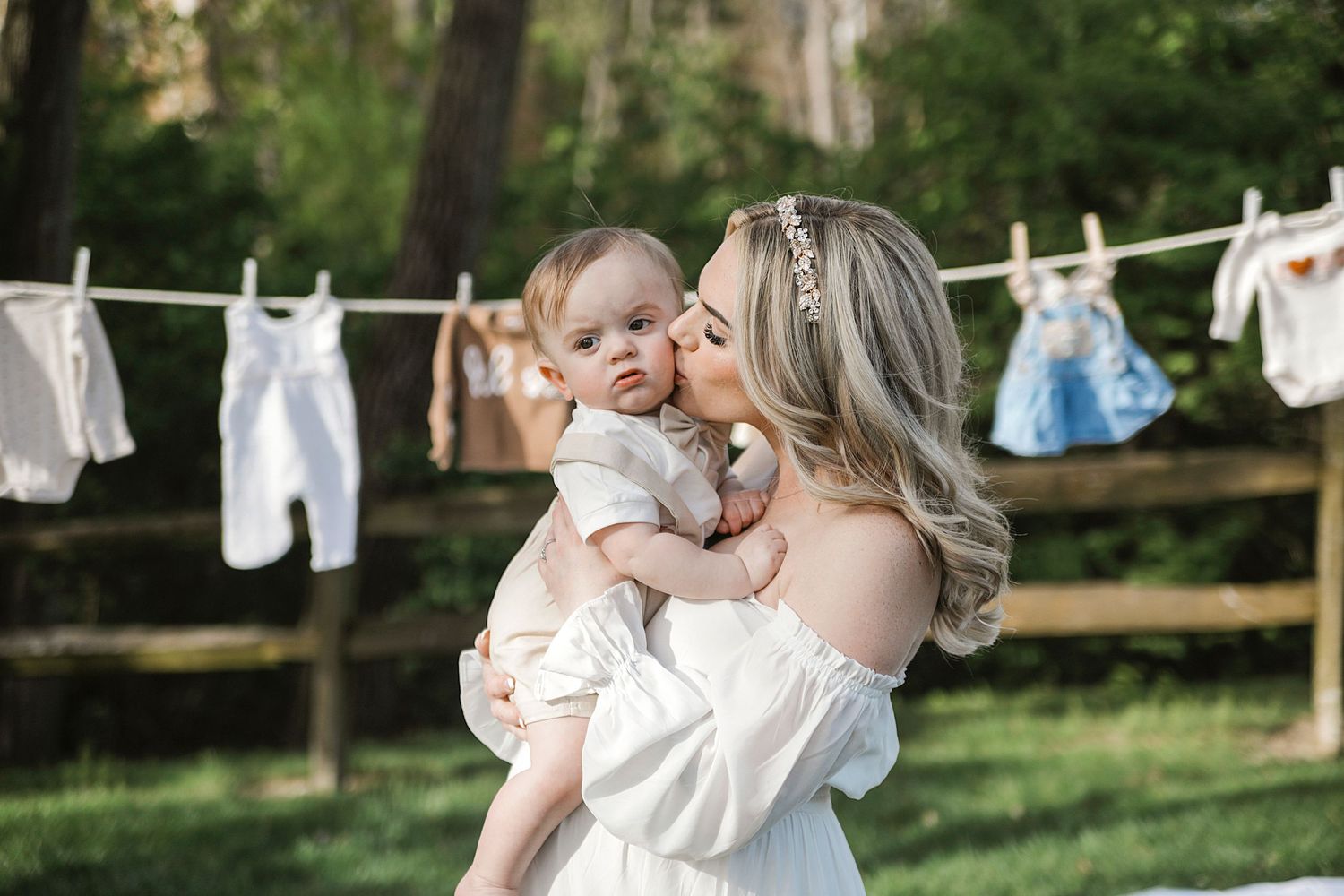 A mother and baby share a tender moment by a rustic clothesline with white laundry hanging in a sunlit backyard.
