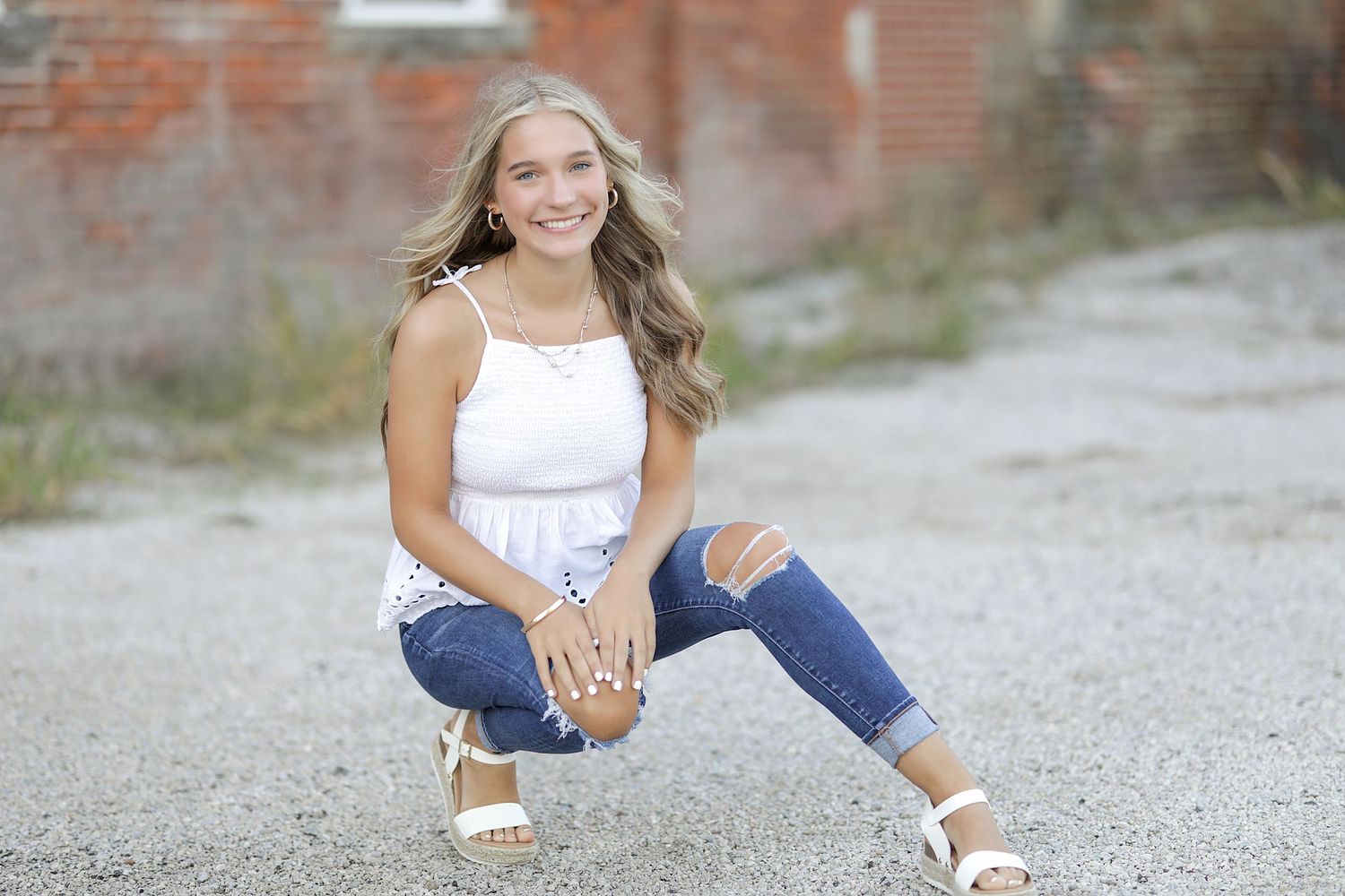 Woman in white tank top and ripped jeans sitting casually on gravel path with brick wall background.
