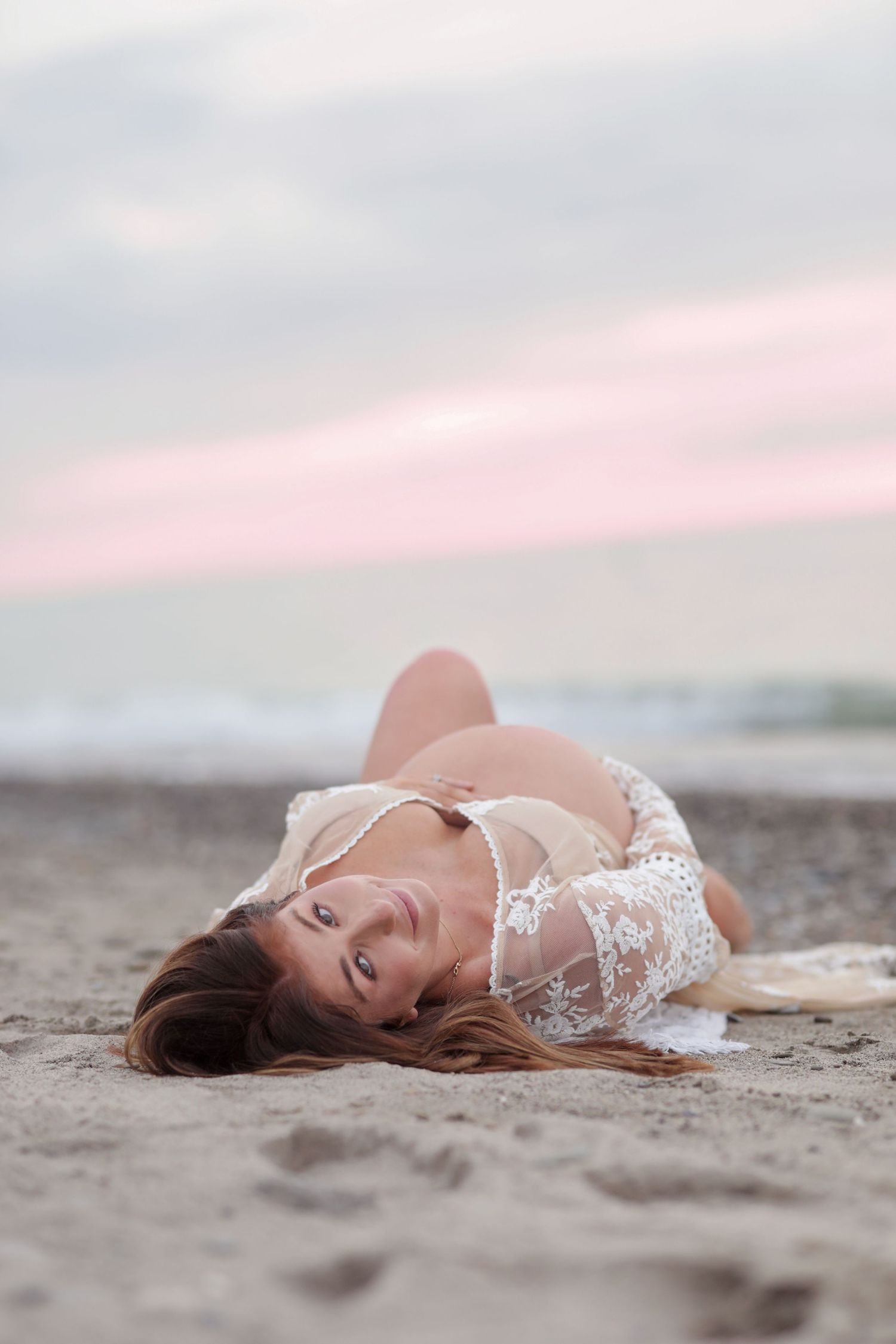 Portrait of someone wearing white lace lying on a beach at sunset with pink and grey sky in background.