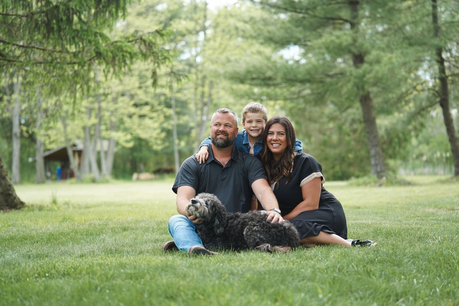 A group sits together on green grass with a large black dog in a wooded outdoor setting.