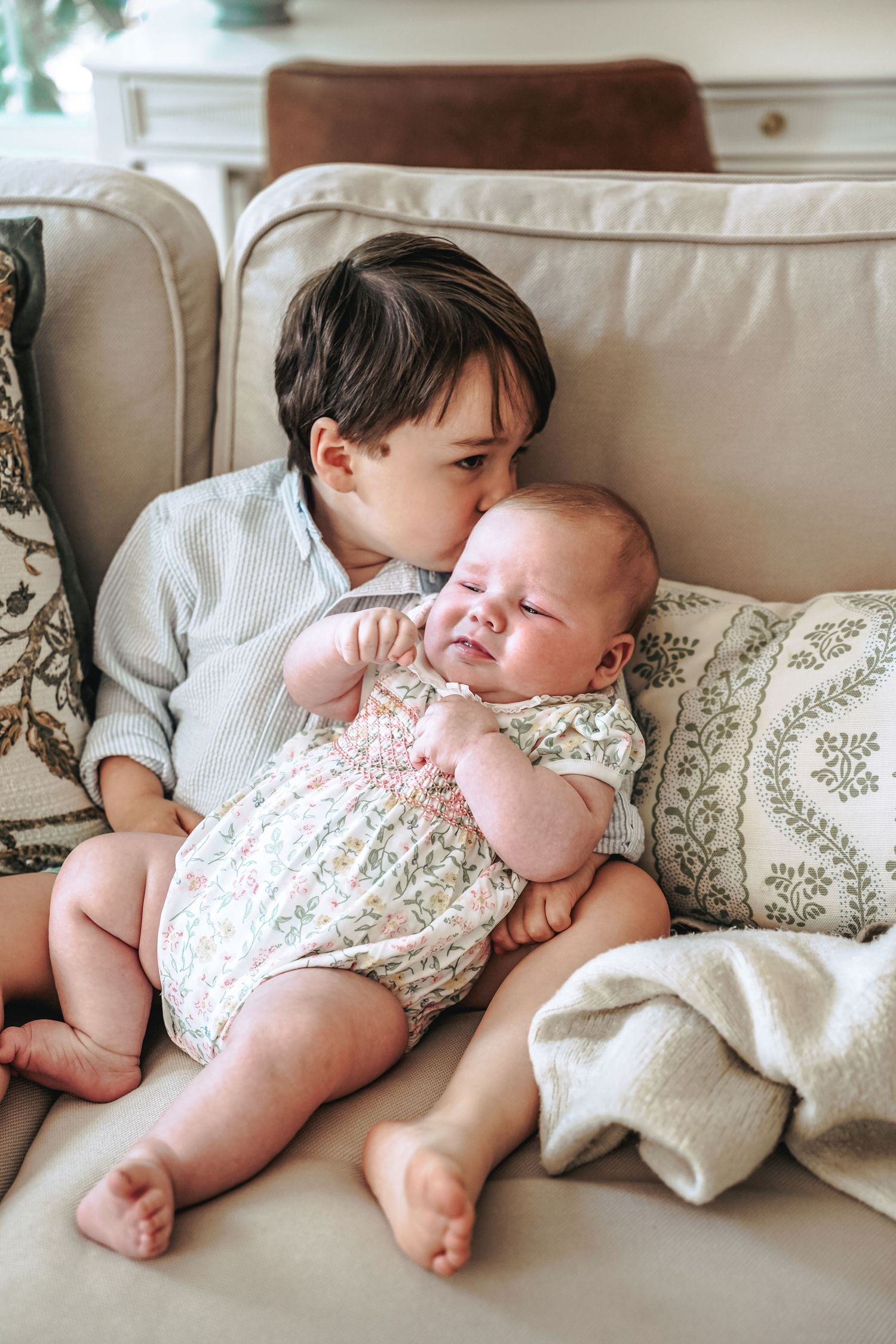 Two siblings share a tender moment together while sitting on a beige couch with decorative throw pillows.