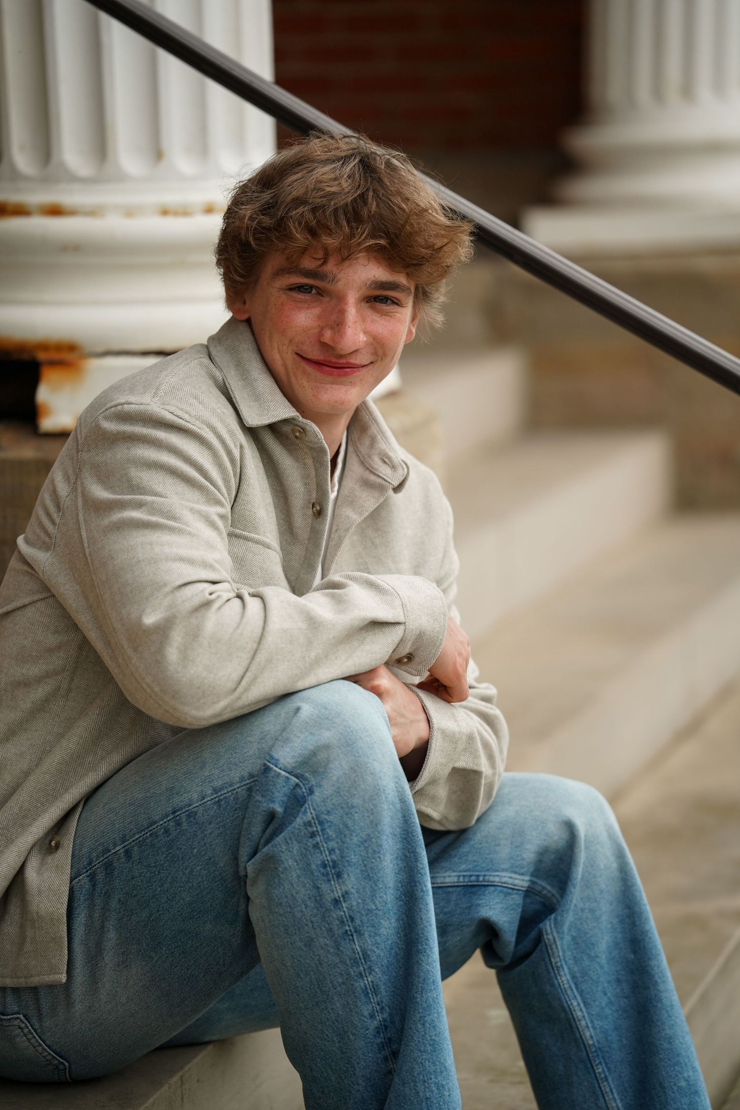 Young person sitting casually on steps wearing a light colored sweater and blue jeans with a column visible in background.