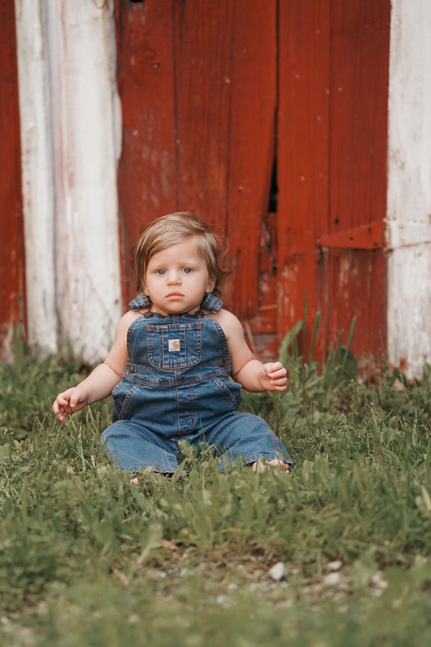 Small child sits in grass wearing denim overalls in front of a rustic red barn door.
