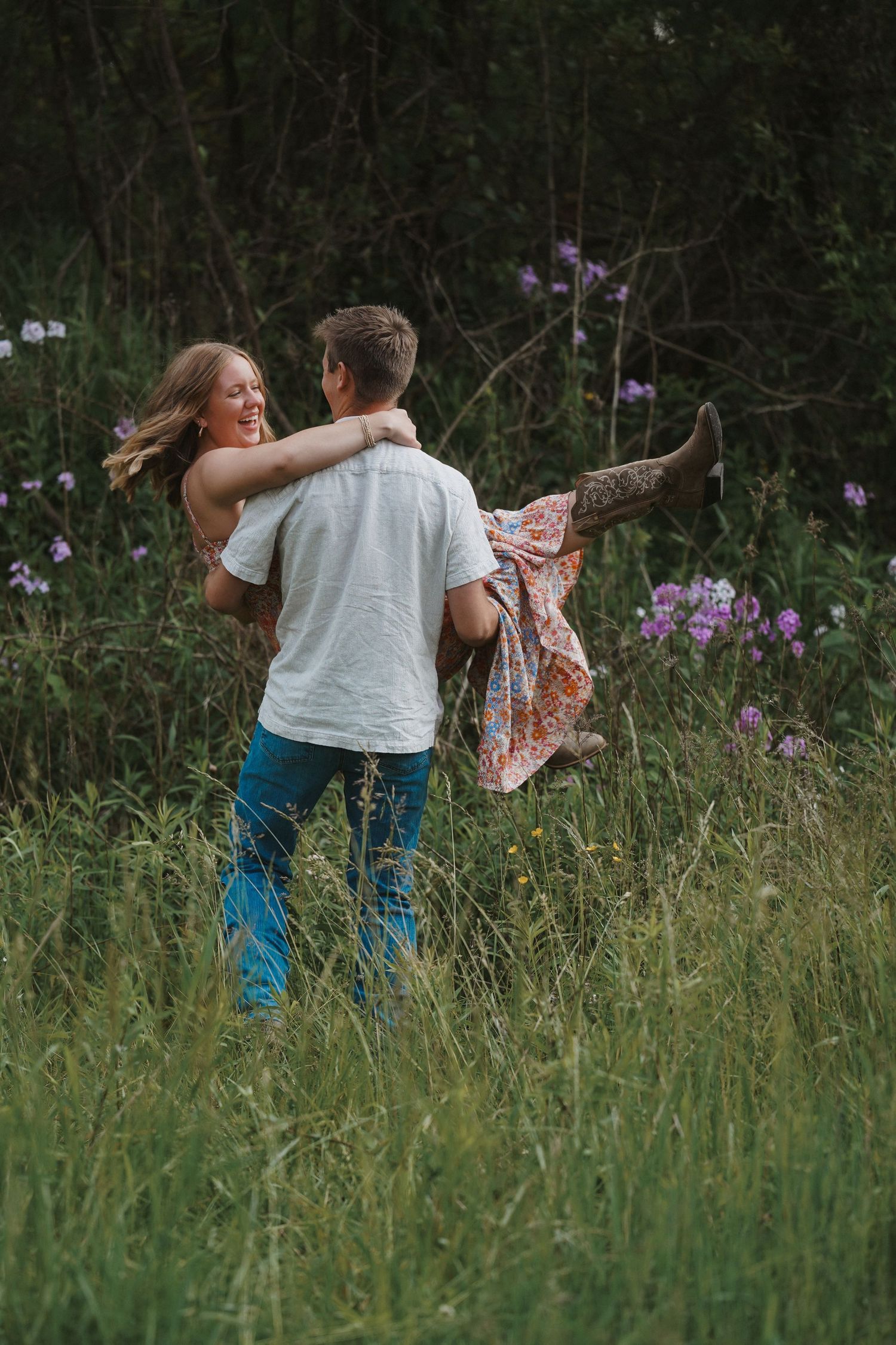 A couple dances and spins playfully in a summer meadow filled with purple wildflowers at dusk.