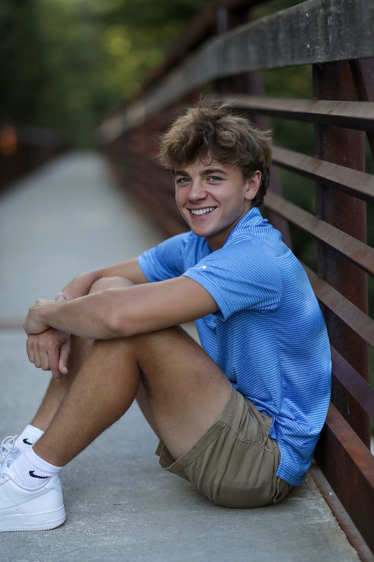 Young person in a blue polo shirt and khaki shorts sits casually against a wooden railing on a bridge or deck.
