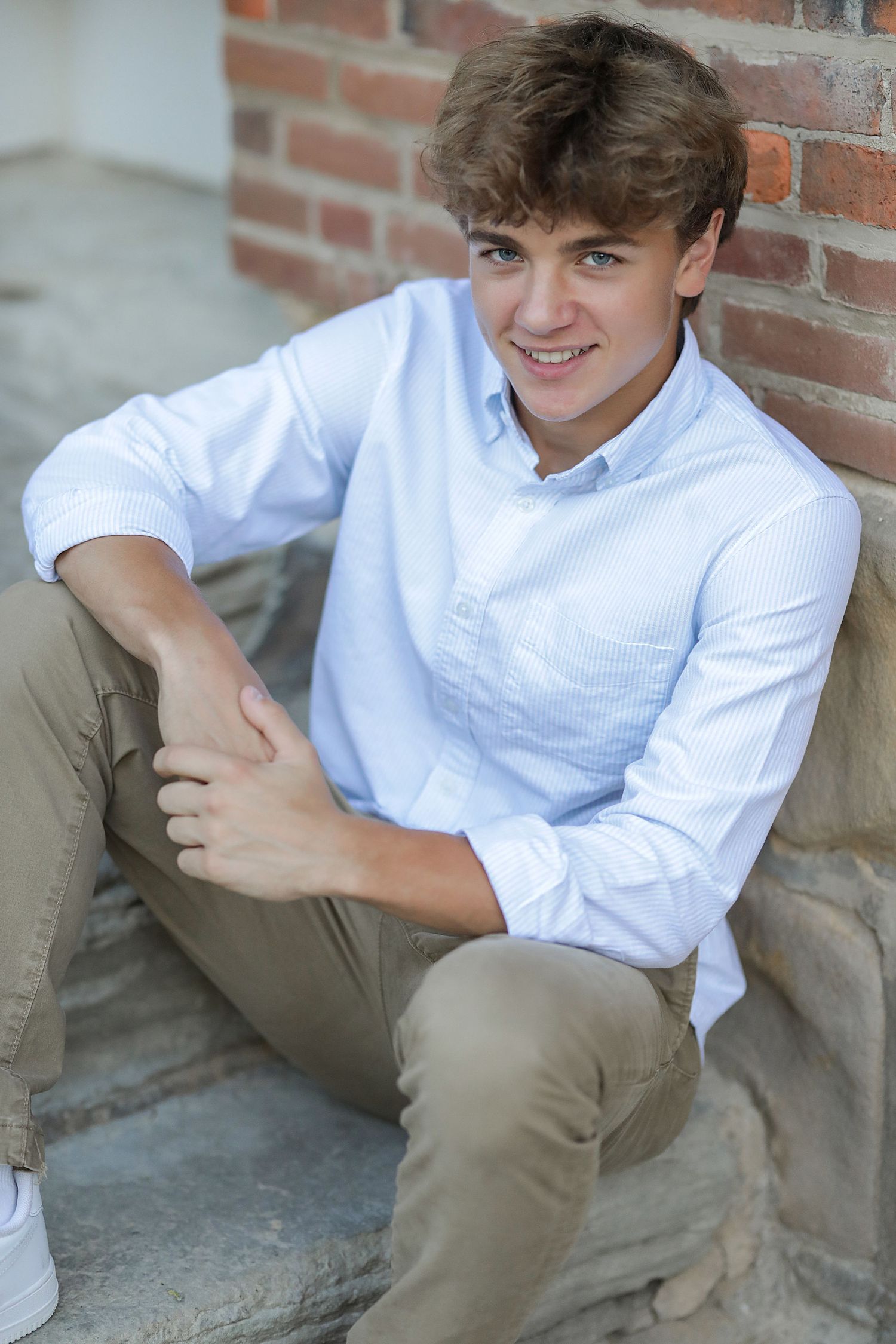 A boy in white button-down shirt and khaki pants sits casually on brick steps during an outdoor portrait session.