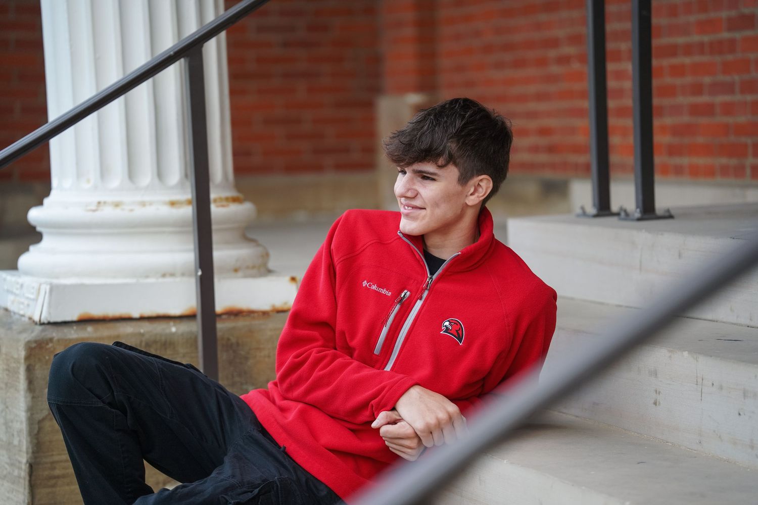 Young student in red pullover sitting on steps against brick wall and white column.