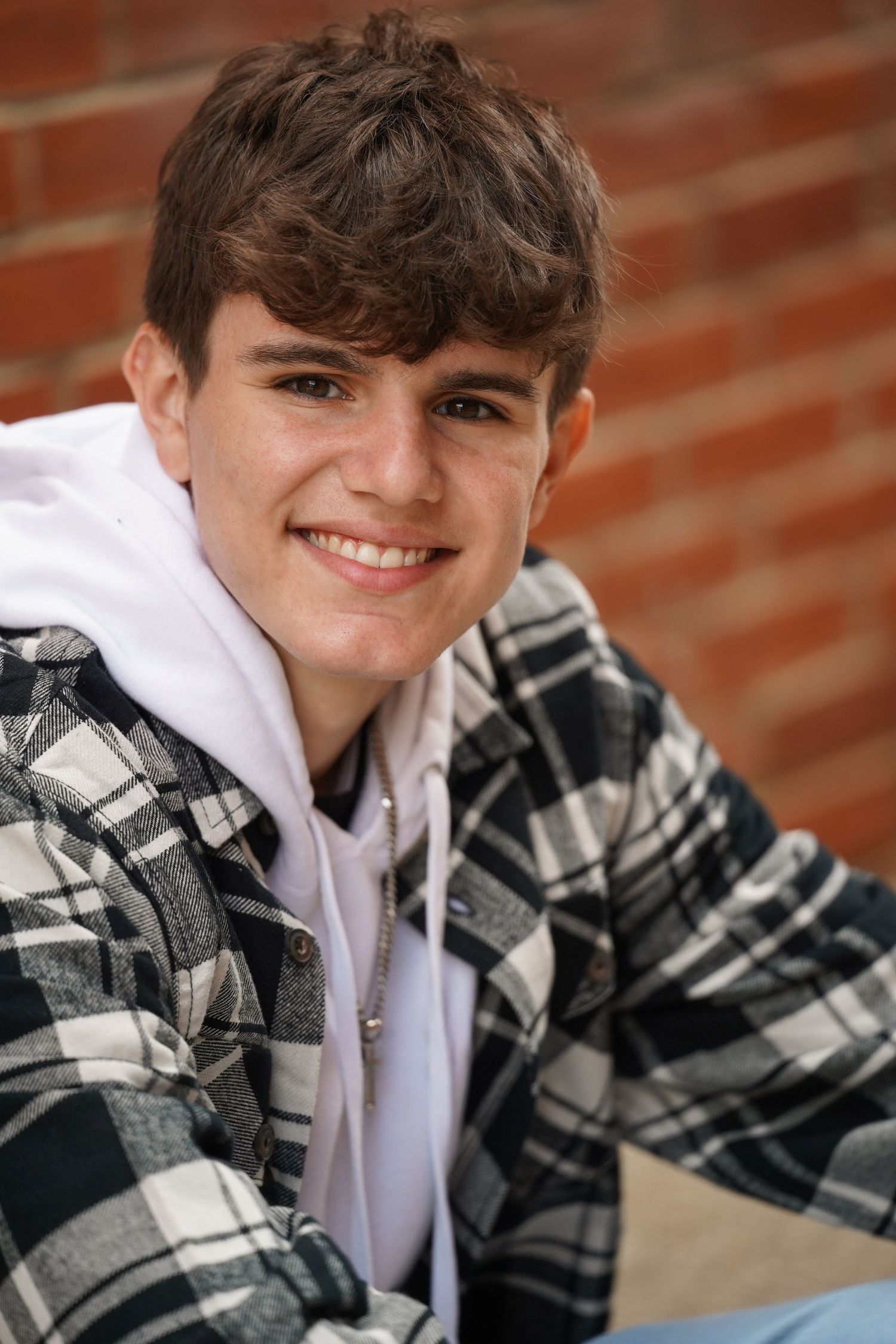 Portrait of someone wearing a plaid flannel jacket over white hoodie against brick wall backdrop.