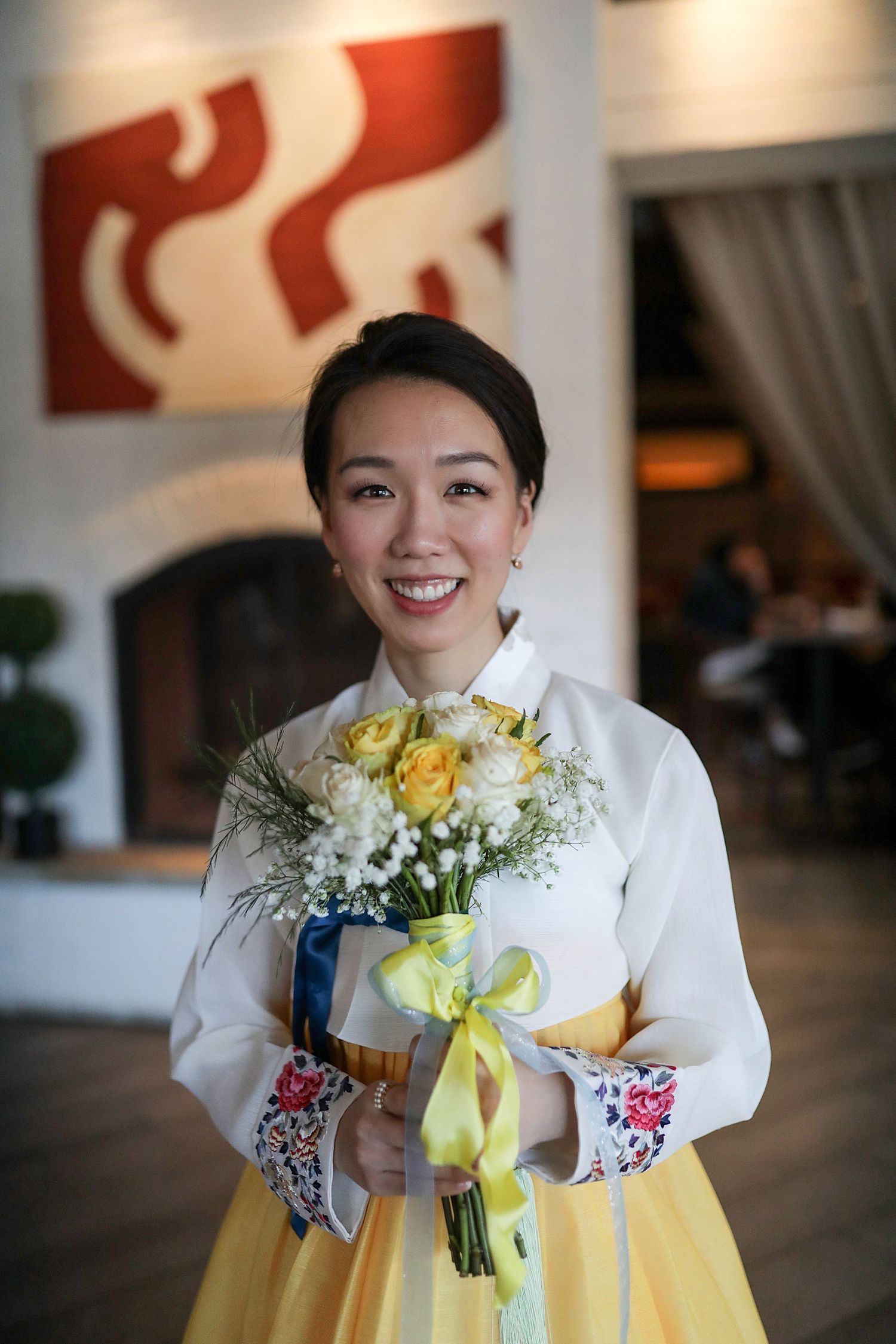 A smiling figure in a traditional Korean hanbok dress holds white and yellow flowers with a yellow ribbon.