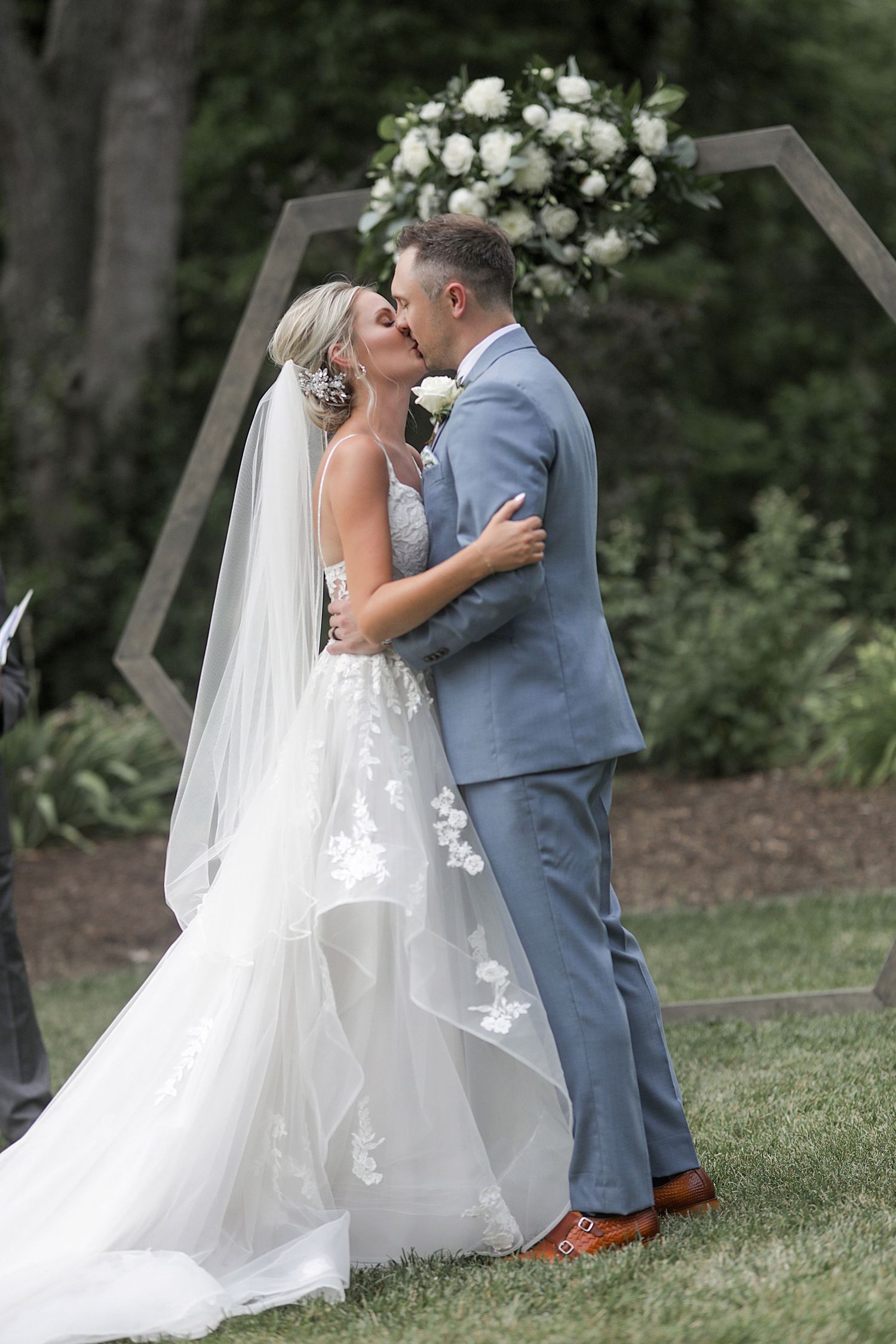 A romantic wedding ceremony moment under a wooden arch decorated with white flowers in an outdoor garden setting.