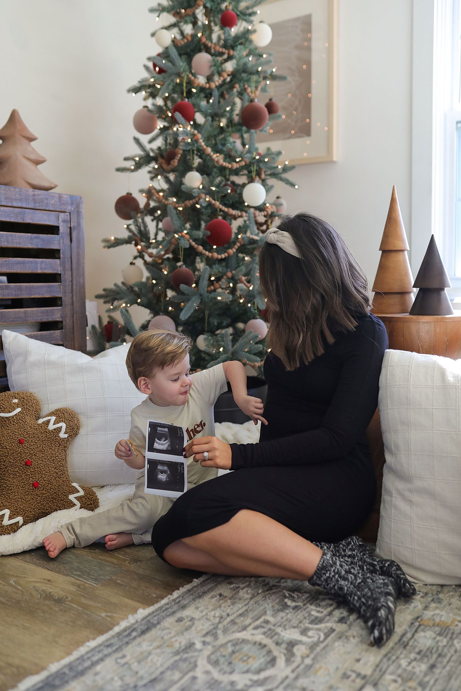 A decorated Christmas tree sits in the corner while two people sit on the floor sharing a special moment near wooden decorations.